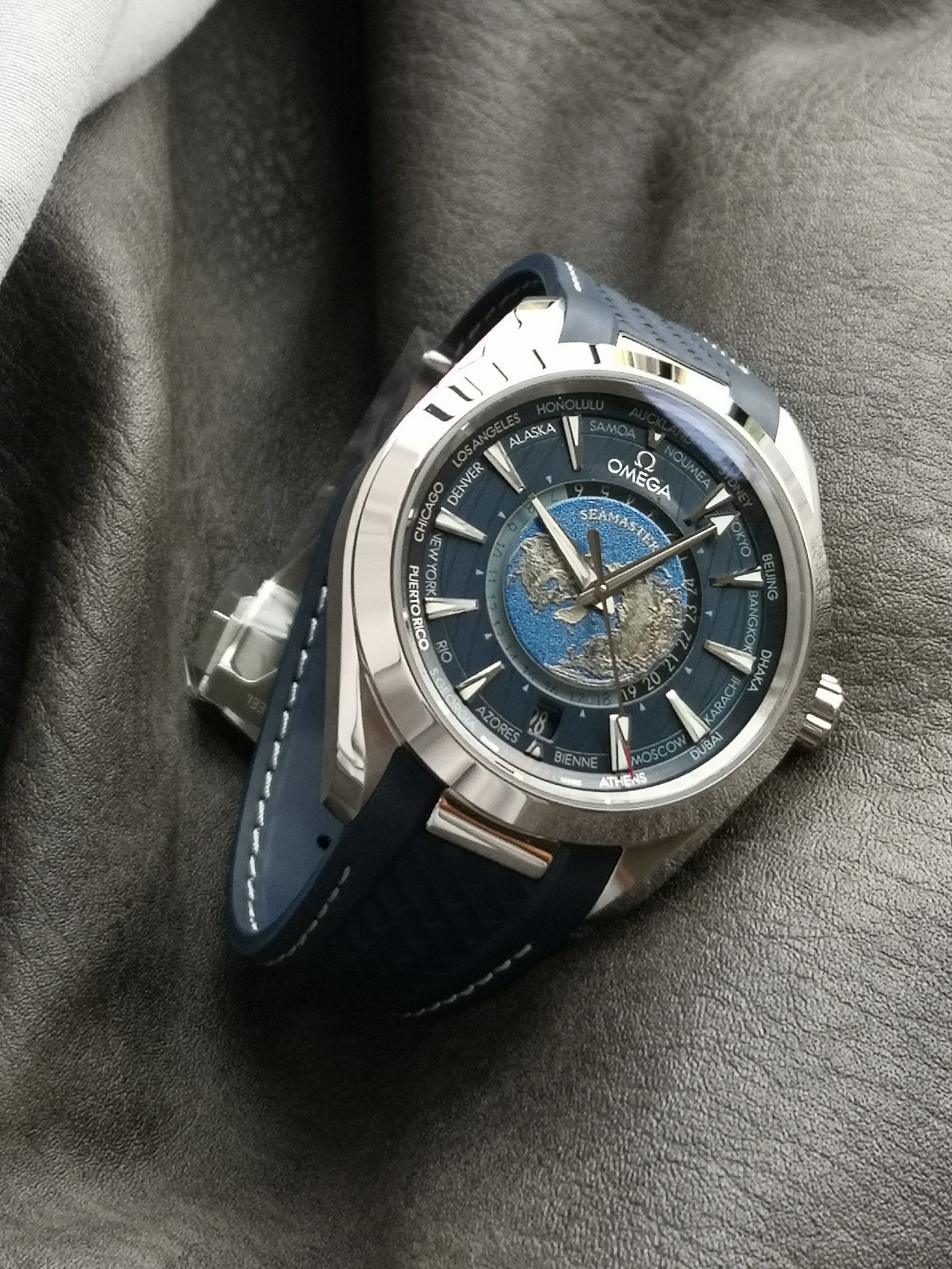 Omega 精钢表壳搭配蓝色天然橡胶表带 蓝色太阳纹饰表盘外侧 饰以垂直柚木纹理 并设有全球目的地时区圈