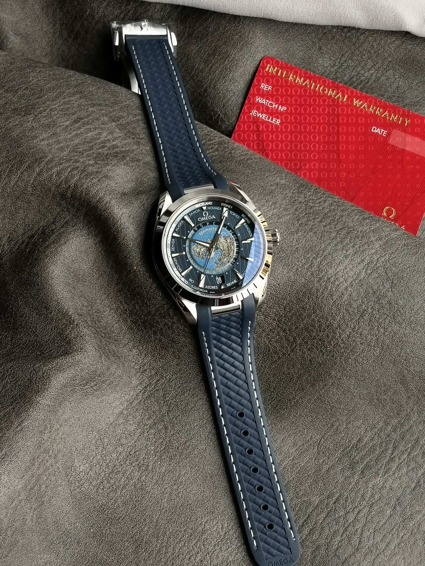 Omega 精钢表壳搭配蓝色天然橡胶表带 蓝色太阳纹饰表盘外侧 饰以垂直柚木纹理 并设有全球目的地时区圈
