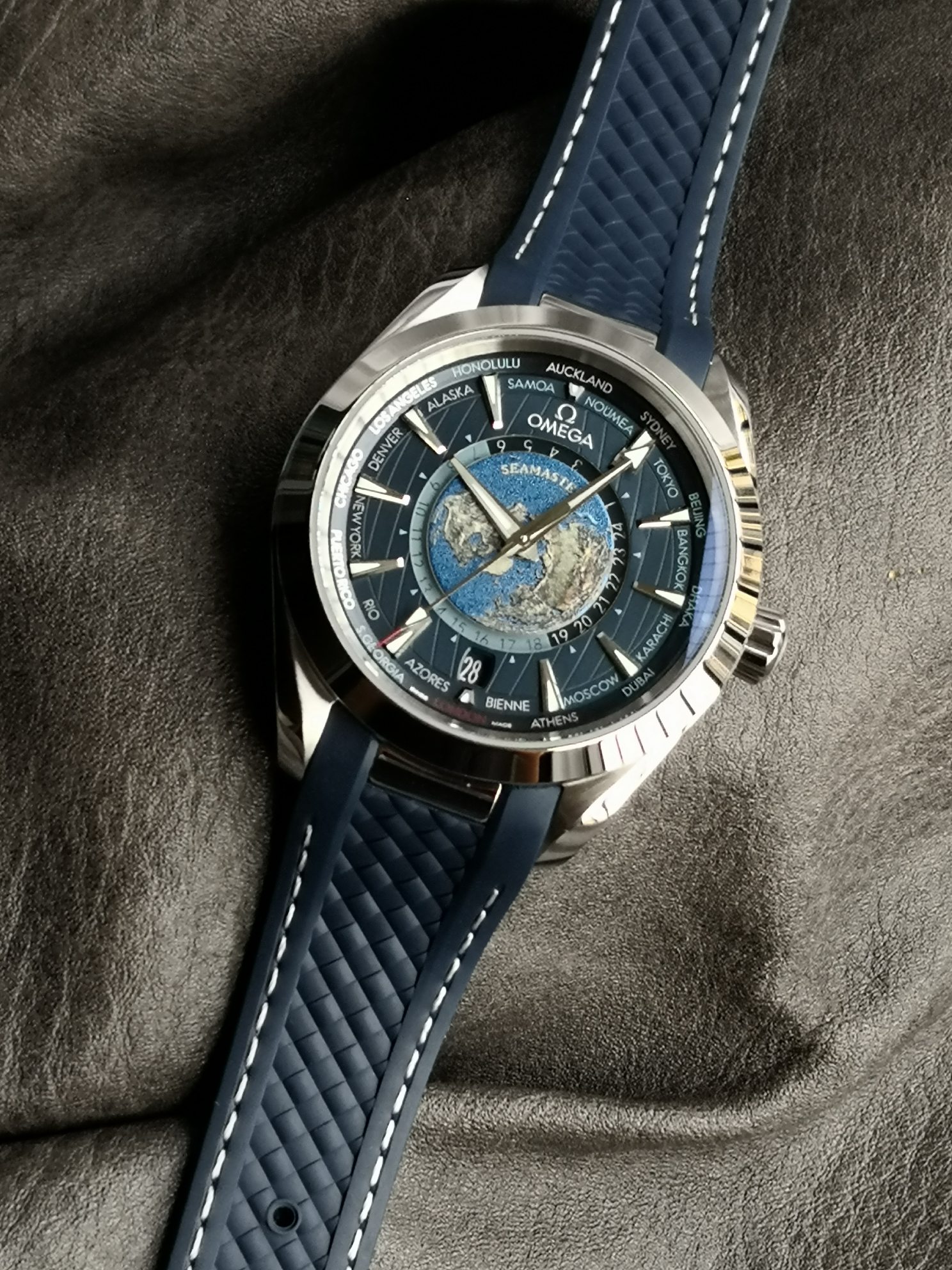 Omega 精钢表壳搭配蓝色天然橡胶表带 蓝色太阳纹饰表盘外侧 饰以垂直柚木纹理 并设有全球目的地时区圈