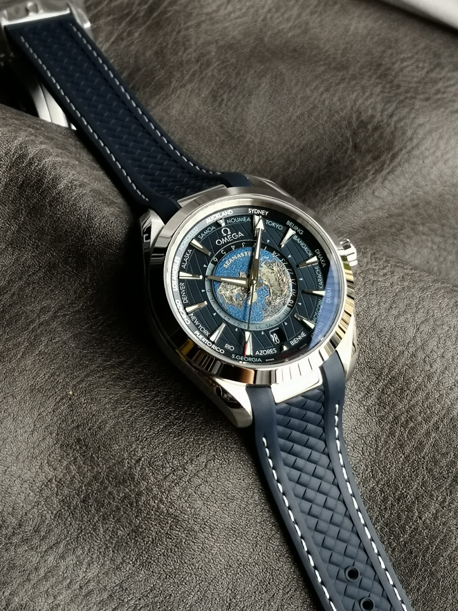 Omega 精钢表壳搭配蓝色天然橡胶表带 蓝色太阳纹饰表盘外侧 饰以垂直柚木纹理 并设有全球目的地时区圈