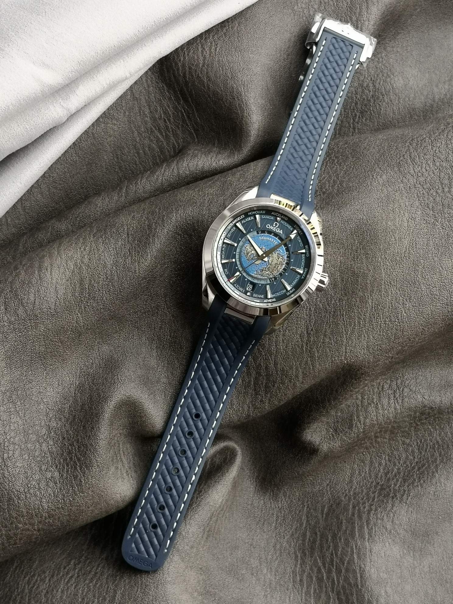 Omega 精钢表壳搭配蓝色天然橡胶表带 蓝色太阳纹饰表盘外侧 饰以垂直柚木纹理 并设有全球目的地时区圈