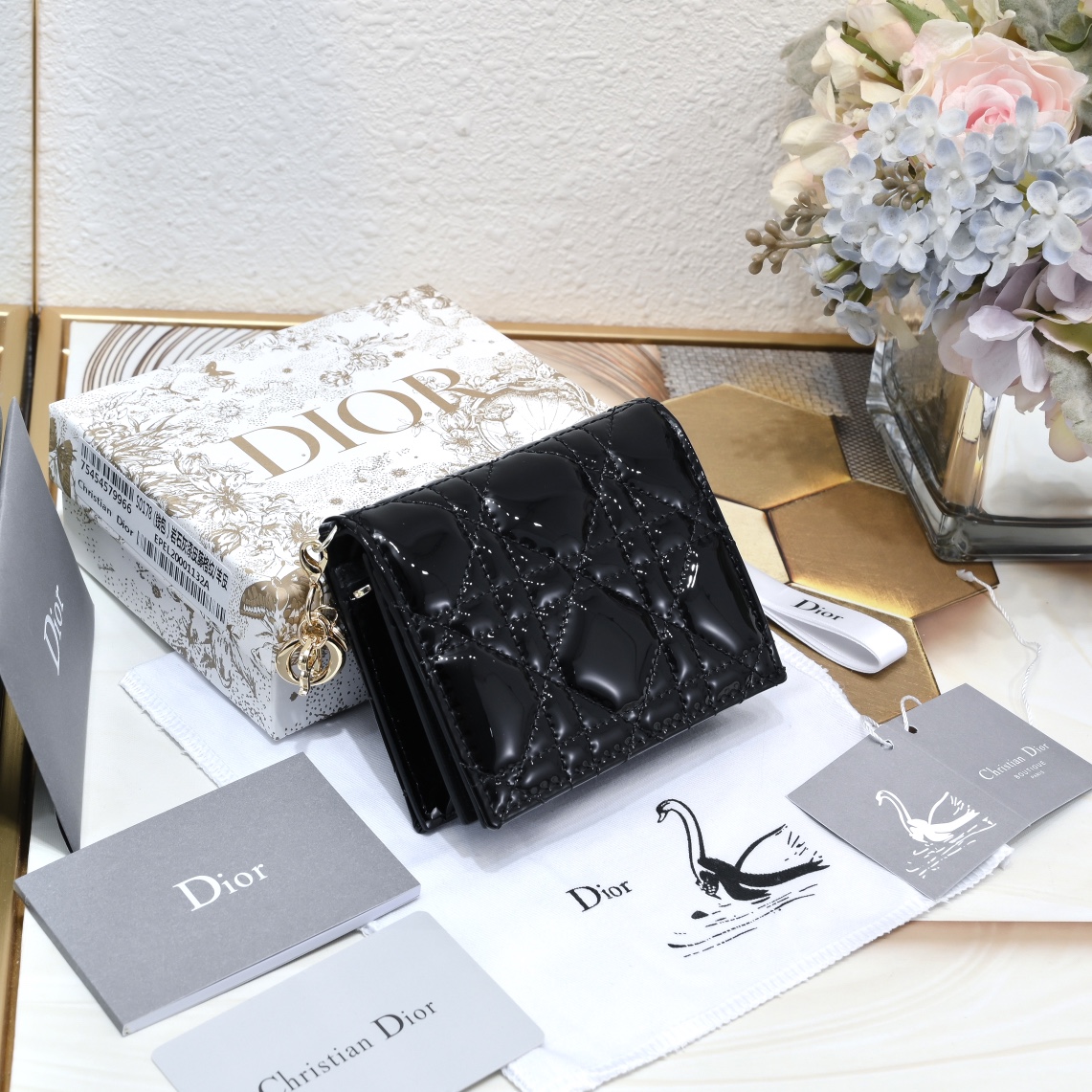 Dior 这款翻盖卡夹采用黑色漆皮牛皮革精心制作，彰显Lady r 系列 设计，搭配藤格纹缉面线和D.I.O.R.吊饰