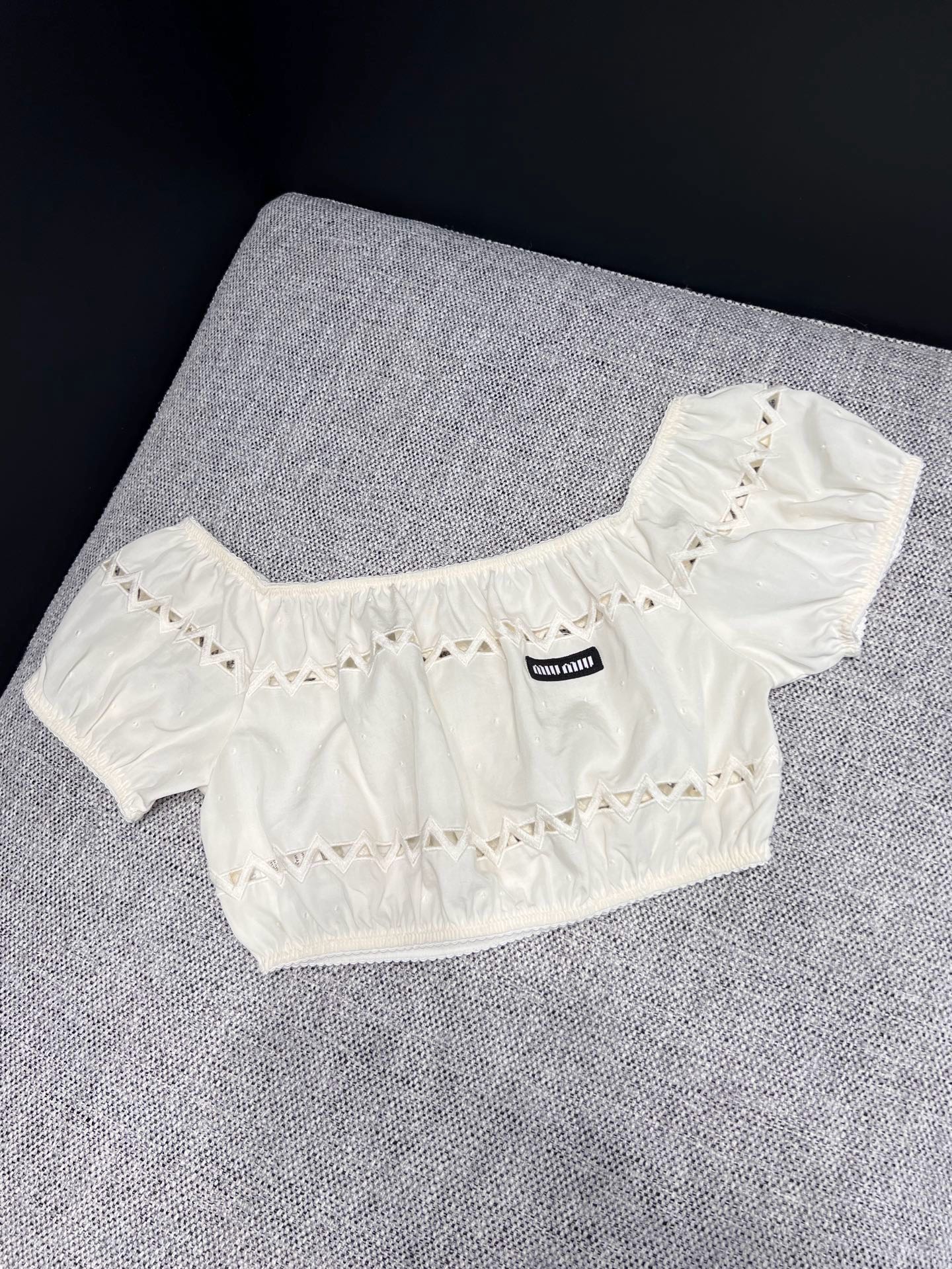 NO:912089,Tops 26 spring and summer new arrivals  Miu** milk white hollow one-shoulder short top, replica of Italian retro sweet temperament. Soft waxy textured cotton material with soft matte texture, elastic one-line collar with puff short sleeves, gently revealing the shoulder and neck lines. The triangular hollow cut all over the body is transparent and light, with exquisite layers and not exaggerated.  The front mini woven label is engraved with the brand logo in a low-key manner, neutralizing the sweetness and adding light luxury. The short waisted version raises the waistline and is suitable for matching with the hollow skirt of the same series. It is suitable for daily appointments and short trips. It is a gentle and smart gold-style top. In stock 36/38/40  Actual detailed pictures,,miumiu,skirts,T-shirt,19860909上衣 26春夏新品 Miu**奶白镂空一字肩短上衣,复刻意式复古清甜气质.软糯肌理棉料自带柔和雾面质感,松紧一字领搭配泡泡短袖,温柔展露肩颈线条.周身错落三角镂空剪裁通透轻盈,层次精致不浮夸,胸前mini织标低调镌刻品牌标识,中和甜腻增添轻奢底蕴.短款收腰版型拉高腰线,适配同系列镂空半裙成套穿搭,日常约会、短途出游皆适配,温柔又灵动的千金风上衣.现货36/38/40 实拍细节图,,miumiu,skirts,T-shirt,,Women's clothing