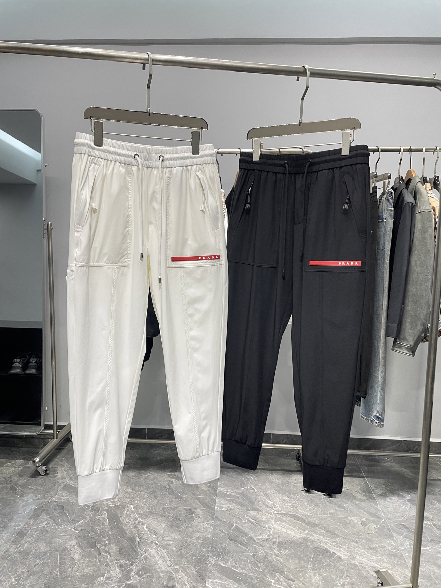 NO:141294,PRA25SS spring and summer new lightweight technology woven drawstring trousers, men's red rubber nameplate fashionable trousers sports and casual trousers cropped trousers!  Trading companies have sparse channels and are on sale on the leading official website!  With its unique fabric selection, exquisite craftsmanship and humanized design details, P home's new hoodies have become an ideal choice for urban people who pursue quality life!  This hoodie uses a customized light and thin technology woven fabric for customer supply. It is not only light and delicate, but also fits like a second layer of skin, but also has excellent breathability and comfort experience!  Even in hot summer or after high-intensity exercise, it can effectively dispel moisture and keep the skin dry and comfortable!  The unique aesthetic concept of the brand is cleverly incorporated into the design. The eye-catching red rubber nameplate decoration under the front pocket is vividly displayed on it, echoing the metal triangle badge on the side of the trouser legs. The two complement each other, not only enhance the overall visual layering, but also inadvertently revealing the extraordinary taste and identity of the wearer, perfectly showing the luxurious style of a high-end brand!  I also put a lot of effort into wearing experience. The waist of the pants adopts a drawstring design, which can freely adjust the tightness. Whether it is a relaxed and comfortable casual style or a tight fit sports experience, it can be easily achieved, meeting the wear needs of different occasions and different needs!  The trouser legs adopt a ribbed splicing tie design. This design not only enhances the wrapping and stability of the trouser, but also makes the trouser shape more three-dimensional and crisp, and is extremely friendly to any leg shape. Whether it is slender and straight long legs or slightly rounded leg lines, they can be perfectly modified to show the best wearing effect!  Color white, black Size M-3XL Size reference M size waist 74 hip circumference 100 thigh circumference 56 thigh circumference 94 L size waist 78 hip circumference 104 thigh circumference 58 thigh circumference 95 XL size waist 82 hip circumference 108 thigh circumference 60 thigh circumference 96 2XL size waist 86 hip circumference 112 thigh circumference 62 thigh circumference 97 3XL size waist circumference 90 thigh circumference 116 thigh circumference 64 thigh circumference 98 08,,prada,alexander wang19860909PRA25SS春夏新款轻薄科技梭织抽绳卫裤,男士红色橡胶铭牌时尚束脚运动休闲长裤九分裤！贸易公司渠道稀出,领先官网发售！P家新款卫裤以其独特的面料选择、精湛的工艺制作以及人性化的设计细节,成为了追求品质生活的都市人群的理想之选！ 这款卫裤采用了客供定制轻薄科技梭织面料,不仅质地轻盈细腻,如同第二层肌肤般贴合,更兼具了卓越的透气性与舒爽体验！即便是在炎炎夏日或是高强度运动后,也能有效排散湿气,保持肌肤的干爽与自在！设计上巧妙地融入了品牌的独特美学理念,前幅口袋下方醒目的红色橡胶铭牌装饰跃然其上,与裤腿侧面金属三角标徽章与之遥相呼应,两者相得益彰,不仅提升了整体的视觉层次感,更在不经意间透露出穿着者不凡的品味与身份,完美展现了作为高端品牌的奢华风范！ 在穿着体验上同样下足了功夫,裤腰部分采用了抽绳式设计,可自由调节松紧度,无论是追求宽松舒适的休闲风格,还是追求紧致贴合的运动体验,都能轻松实现,满足了不同场合、不同需求的穿着需求！而裤脚则采用了罗纹拼接束口设计,这一设计不仅增强了裤装的包裹性与稳定性,更使得裤型更加立体挺括,对任何腿型都极为友好,无论是纤细笔直的长腿,还是略显圆润的腿部线条,都能被完美修饰,展现出最佳的穿着效果！ 颜色白色、黑色 尺码M-3XL 尺寸参考 M码腰围74臀围100大腿围56裤长94 L码腰围78臀围104大腿围58裤长95 XL码腰围82臀围108大腿围60裤长96 2XL码腰围86臀围112大腿围62裤长97 3XL码腰围90臀围116大腿围64裤长98 08,,prada,alexander wang,Men's clothing