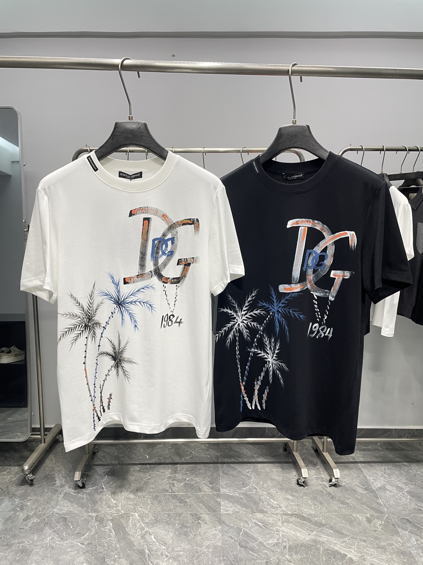 NO:857895,DG26SS new spring and summer coconut tree graffiti printed cotton round neck short-sleeved T-shirt!  A perfect fusion of Italian romance and street art, this T-shirt is Dolce & Gabbana's modern interpretation of summer style. It is made of combed cotton fabric, which is soft to the touch and skin-friendly, has excellent breathability and perspiration performance, and is not easy to deform after long-term wear. The inside of the collar is decorated with a brand webbing label to enhance recognition; the body printing uses environmentally friendly digital direct printing technology, which has high color saturation and is durable and washable. The delicate graffiti effect restores the hand-painted texture!  With the palm tree as the core visual element, paired with the DG logo with aged strokes and the 1984 year number, it cleverly combines the southern Italian resort style with the brand's founding year. Available in black and white, it not only retains the brand's iconic sense of luxury, but also injects a casual and uninhibited street art atmosphere. It is an ideal choice for modern men to show their individual taste.  The white model can be paired with light blue denim shorts and woven sandals to create a refreshing holiday atmosphere; the black model can be paired with overalls and Martin boots to easily create a cool street look; it can also be worn with a light linen shirt to expose the printed details on the chest, adding an artistic touch to the business casual style and suitable for daily commuting, holiday travel and other diverse scenes. Color white, black Size S-XXL Size reference S size shoulder width 43 bust 100 length 69 M size shoulder width 44 chest 104 length 70  L size shoulder width 45 bust 108 length 71 XL size shoulder width 46 bust 112 length 72 219860909DG26SS春夏新款椰树涂鸦印花纯棉圆领短袖T恤！ 将意式浪漫与街头艺术完美融合,这件T恤是杜嘉班纳对夏日风情的摩登诠释. 采用精梳棉面料,触感柔软亲肤,透气排汗性能优异,久穿不易变形.领口内侧饰有品牌织带标签,强化辨识度；衣身印花采用环保数码直喷工艺,色彩饱和度高且牢固耐洗,笔触细腻的涂鸦效果还原手绘质感！以棕榈椰树为核心视觉元素,搭配做旧笔触的DG标识与1984年份数字,将南意度假风情与品牌创始年份巧妙结合,黑白两色可选,既保留了品牌标志性的奢华感,又注入了随性不羁的街头艺术气息,是现代男士彰显个性品味的理想之选. 白色款可搭配浅蓝牛仔短裤与编织凉鞋,营造清爽度假氛围；黑色款搭配工装裤与马丁靴,轻松打造酷感街头造型；亦可外搭轻薄亚麻衬衫,露出胸前印花细节,为商务休闲风增添艺术感,适配日常通勤、假日出游等多元场景. 颜色白色、黑色 尺码S-XXL 尺寸参考 S码肩宽43胸围100衣长69 M码肩宽44胸围104衣长70 L码肩宽45胸围108衣长71 XL码肩宽46胸围112衣长72 2XL码肩宽47胸围116衣长73,,Dolce&Gabbana,sandals,boots,shorts,shirts,T-shirt,tees，t-shirt,,Men's clothing