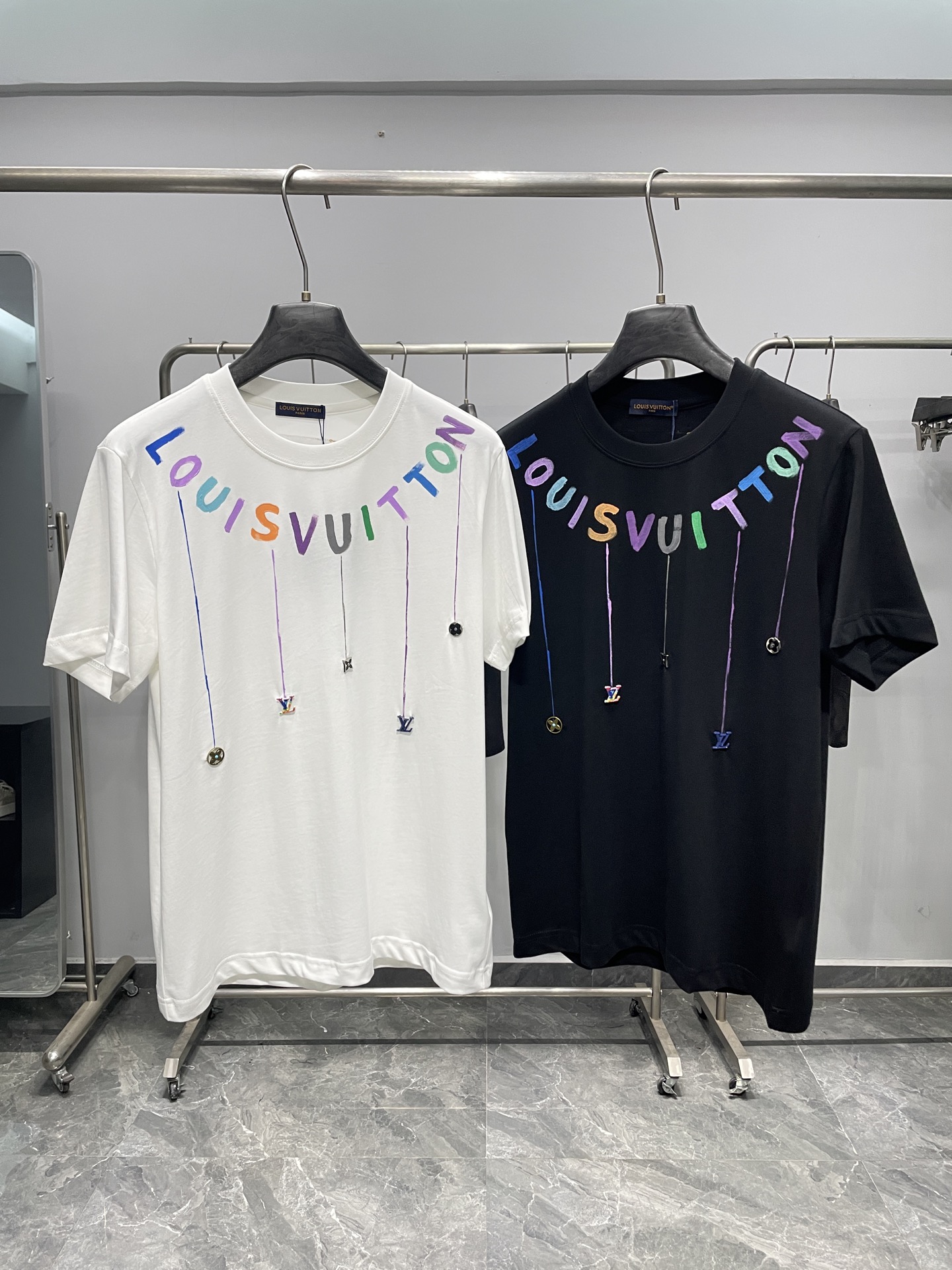 NO:857884,1V26SS new spring and summer colorful hand-painted letter pendant pure cotton round neck short-sleeved T-shirt!  This short-sleeved T-shirt combines artistic hand-painting with three-dimensional accessories, giving the brand logo a lively and trendy feel.  High-count combed cotton fabric is selected, pre-shrunk and anti-wrinkle treated, and feels soft and skin-friendly, with both drape and stiffness. The fine knitted texture brings a warm texture and is breathable and sweat-wicking. It is suitable for daily wear in spring and summer. It is not easy to deform and pill after long-term wear, taking into account both comfort and durability. The classic round-neck version, neat tailoring modifies the body shape, and does not care about body proportions. The body of the garment uses colorful hand-painted letters arranged in an arc as the core visual, blue  , purple, green, orange and other multi-colored brushstrokes create a casual artistic feel; multiple pendant lines extend below the letters, connecting the 1V logo, Monogram flowers, diamond patterns and other three-dimensional metal pendants. The colors and materials are rich in layers, low-key highlighting the brand style. A dark blue logo label is sewn on the inside of the collar, and a reverse patch label on the back collar further enhances the sense of luxury. Black and white are available to meet different style needs.  The overall style leans towards the light luxury street art style, which not only retains the elegance of Louis Vuitton, but also incorporates the fun sense of modern trends. The white model is paired with light-colored denim shorts and white shoes to create a fresh and energetic street look. The black model is paired with functional overalls and Martin boots to release a cool and uninhibited street atmosphere. Whether worn alone or as a layered jacket, it can easily be used in daily outings, trendy gatherings and other diverse scenes. It is a luxury basic style in the wardrobe that is both recognizable and interesting.  Color White, Black Size M-3XL Size Reference M Size Shoulder Width 43 Bust 100 Length 69 L Size Shoulder Width 44 Bust 104 Length 70  & coats,T-shirt,tees,t-shirt,198609091V26SS春夏新款彩色手绘字母吊坠纯棉圆领短袖T恤！ 这款短袖T恤以艺术手绘与立体配饰的碰撞,将品牌标识玩转出鲜活潮流感. 甄选高支精梳棉面料,经过预缩与抗皱处理,触感柔软亲肤,垂坠感与挺括度兼备.细密针织纹理带来温润质感,透气排汗的天然属性,适配春夏日常穿搭,久穿不易变形起球,兼顾舒适与耐穿性.经典圆领版型,利落剪裁修饰身形,不挑身材比例.衣身以弧形排列的彩色手绘字母为核心视觉,蓝、紫、青、橙等多色笔触营造随性艺术感；字母下方延伸出多条垂坠线条,串联起1V标识、Monogram花卉、菱形纹等立体金属吊坠,色彩与材质层次丰富,低调彰显品牌格调.领口内侧缝制深蓝色标识标签,后领处的反向贴布标签进一步强化奢品质感,黑白两色可选,满足不同风格需求. 整体风格偏向轻奢街头艺术风,既保留了路易威登的优雅底蕴,又融入了现代潮流的玩趣感,白色款搭配浅色牛仔短裤与小白鞋可打造清新活力的出街造型,黑色款搭配机能工装裤与马丁靴则能释放酷飒不羁的街头氛围,无论是单穿还是作为内搭叠穿外套,都能轻松驾驭日常出街、潮流聚会等多元场景,是衣橱中兼具辨识度与趣味性的奢品基础款. 颜色白色、黑色 尺码M-3XL 尺寸参考 M码肩宽43胸围100衣长69 L码肩宽44胸围104衣长70 XL码肩宽45胸围108衣长71 2XL码肩宽46胸围112衣长72 3XL码肩宽47胸围116衣长73,,louis vuitton,boots,shorts,jackets & coats,T-shirt,tees，t-shirt,,Men's clothing
