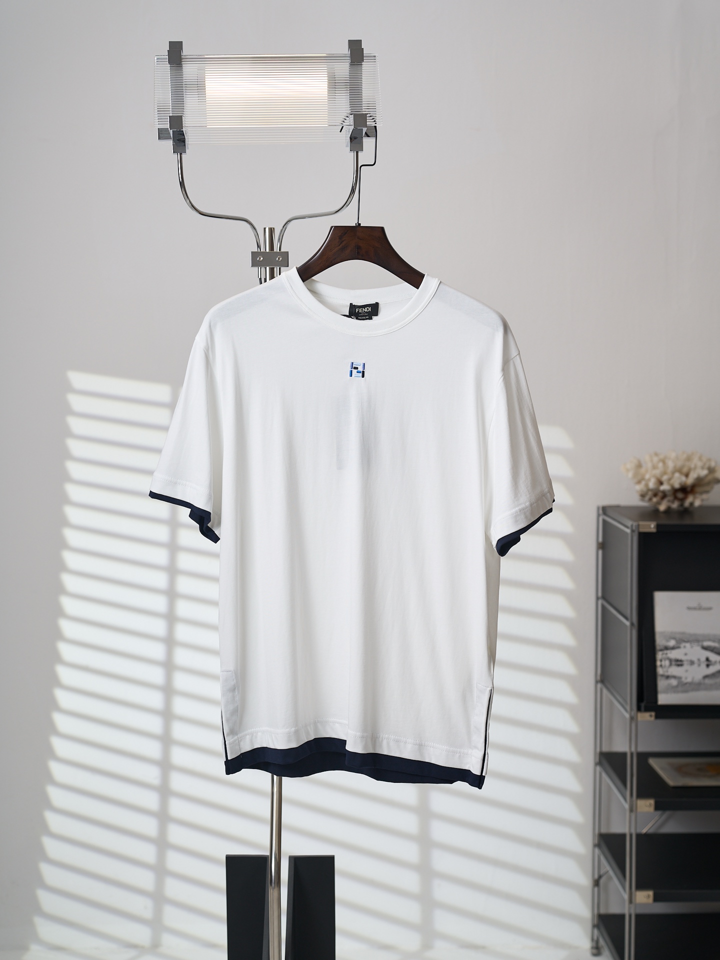 NO:931808,[Real auction] FENDI/Fendi FF color embroidered fake two-piece round neck short-sleeved T-shirt [Ready stock] XS S M L fabric 24 count double-sided plain weave / 250G rib 24 count 1×1 rib / 300G  It uses 24-count double-sided pure cotton plain knitted fabric. The double-sided knitted structure is woven with two layers of coils intertwined with each other. The front and back sides have the same appearance, giving the fabric the stability and double thickness of the same pattern on the front and back. It not only retains the skin-friendly breathability of pure cotton, but also has a crisp and skeletal feel.  and excellent drape. Since this model is a fake two-piece design, the white body and the dark blue contrasting trim need to be accurately aligned and stitched. The color difference is large, and the stitching must be tight and smooth; ensure that the edges of the fake two-layer structure are clean and neat, showing a seamless layering effect.  The blue combined FF logo on the chest is the soul of this T-shirt's craftsmanship. The classic intertwined double F letters have a slender structure and complex twists, which require extremely high embroidery precision. The two-color embroidery uses both dark blue and light blue embroidery threads, and adopts a flat stitch method - filling the pattern area with high-density, parallel-arranged linear stitches to form a smooth, flat, and full embroidery surface with uniform color and strong coverage.  In order to enhance the three-dimensional sense, the process increases the number of embroidery thread layers and uses thicker embroidery threads to make the pattern clearly protrude from the surface of the fabric, presenting a touchable relief feeling. The final effect is an exquisite brand logo with clear outlines, full colors, and a significant three-dimensional convex feel. It has a solid touch and a high sense of quality visually.,,fendi,fendi,T-shirt,tees,t-shirt,19860909【实物挂拍】FENDI/芬迪 FF彩色刺绣假两件圆领短袖T恤【现货】XS S M L 面料24支双面平纹 / 250G 罗纹24支1×1罗纹 / 300G 选用24支双面纯棉平纹针织面料,双面针织结构通过两层线圈相互串套编织,正反面外观一致,赋予面料正反同纹的稳定性与双倍厚度,既保留纯棉的亲肤透气性,又兼具挺括的骨感与优异的垂坠度.由于本款为假两件设计,白色大身与深蓝色撞色饰边需要精确对位缝合,颜色差异大,缝线必须紧实平整；确保假两层结构的边缘干净利落,呈现浑然一体的叠穿效果. 胸前蓝色系组合FF标志是该款T恤的工艺灵魂所在.经典交织双F字母结构纤细、转折繁复,对刺绣精密度要求极高.双色刺绣同时使用深蓝与浅蓝绣线,采用平针针法——通过高密度、平行排列的直线针脚填充图案区域,形成光滑、平整、饱满的绣面,色彩均匀且覆盖力强. 为强化立体感,工艺中通过增加绣线层数并使用较粗绣线,使图案明显凸出于面料表面,呈现触手可及的浮雕感.最终效果是轮廓清晰、色彩饱满、具有显著立体凸起感的精致品牌标识,触感扎实,视觉上极具品质感.,,fendi,fendi,T-shirt,tees，t-shirt,,Men's clothing
