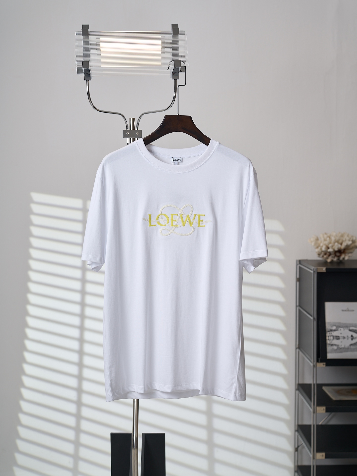 NO:931775,[Physical hanging auction] LOEWE/Loewei two-color logo hemp rope embroidered cotton round neck short-sleeved T-shirt [ready stock] XS S M L fabric 24 count special woven compact cotton / 285G rib 32 count 1×1 rib / 340G This short-sleeved shirt is made of 24-count special-spun compact cotton, which is a medium-thick knitted fabric that is both stiff and comfortable. The compact spinning technology makes the fibers arranged parallel, hairiness is significantly reduced, and the surface is smooth and smooth. After shaping, the fabric has both stiffness and softness, and a moderate drape, which provides a smooth and stable base fabric for subsequent two-color embroidery. Embroidery uses hemp rope as the core material, and combines two techniques: tatami embroidery and punch stitch. Tatami embroidery uses closely arranged parallel stitches to form a flat surface like a mat, giving the rough natural texture of the hemp rope a sense of order; punch stitch uses short overlapping stitches to outline the outline of the letters, adding a smart layer. The rustic texture of hemp rope and the two embroidery methods combine hardness and softness - tatami embroidery solidifies the base and shows the sincerity and strength; the stitching details balance the overall rhythm. The letter logo itself adopts stitching embroidery technology. The embroidery needles are staggered back and forth as they move, forming a fine and rhythmic line texture, which is as vivid as hand-painted brushstrokes. The three-dimensional effect of light and shadow flow is created through changes in angle and density.,,loewe,loewe,T-shirt,tees,t-shirt,19860909【实物挂拍】LOEWE/罗意威 双色标识麻绳刺绣棉质圆领短袖T恤【现货】XS S M L 面料24支特纺紧密棉 / 285G 罗纹32支1×1罗纹 / 340G 这款短袖选用24支特纺紧密棉,属于兼具挺括与舒适的中厚型针织面料.紧密纺技术使纤维排列平行、毛羽显著减少,表面光洁平滑.定形后的面料兼具挺括性与柔软度,垂坠感适中,为后续的双色刺绣提供了平整稳定的底布基础. 刺绣以麻绳为核心材质,融合塌塌米绣与挨针两种技法.塌塌米绣通过紧密排列的平行针脚,形成平整如席的块面,赋予麻绳粗犷的天然肌理以秩序感；挨针则以短促交叠的针迹勾勒字母轮廓,增添灵动层次. 麻绳的质朴质感与两种绣法刚柔相济——塌塌米绣夯实基底,展现朴拙力道；挨针细节平衡整体节奏.字母标识本身采用挨针刺绣工艺,绣针行进中前后错落,形成细密且富有节奏感的线条肌理,犹如手绘笔触般生动,通过角度与疏密变化塑造出光影流动的立体效果.,,loewe,loewe,T-shirt,tees，t-shirt,,Men's clothing