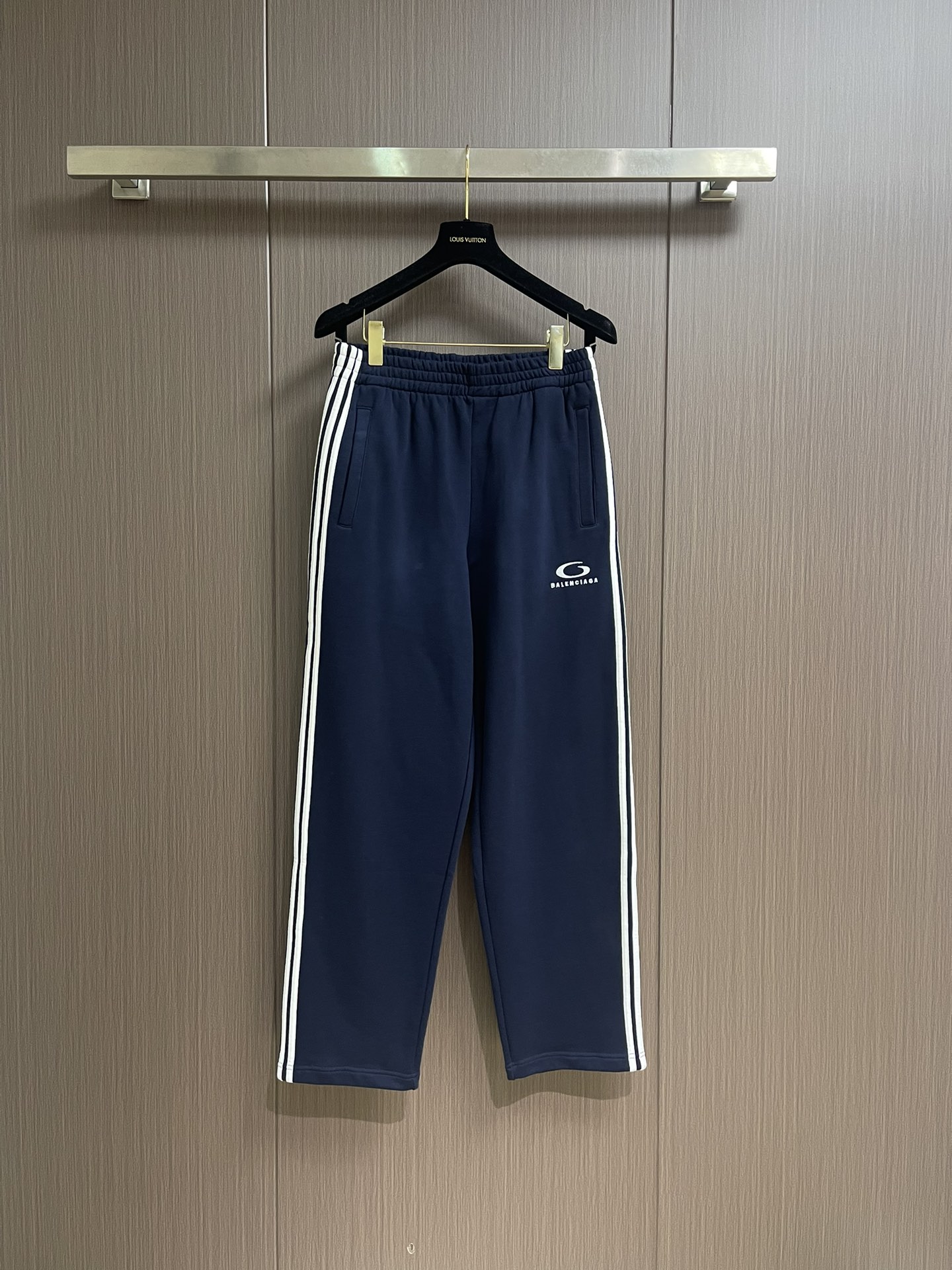 NO:524770,BALENCIAGA embroidered sweatpants are made of single-sided fish scale fabric woven with cotton as the base. The front is a smooth knitted surface. The touching surface is formed by the floating thread knitting technology of the knitting machine to form a three-dimensional loop, which increases the fluffy feeling and is delicate at the same time. The loop structure absorbs a large amount of water and evaporates quickly, and it is not easy to deform loosely after washing multiple times. The imported embroidery machine is 3000 stitches + decorated with loop sports icon embroidery on the front side of the thigh. The double-axis linear guide drives the embroidery frame, and the frame shift accuracy can reach 0.01mmP, ensuring the precise positioning of the embroidery pattern. The decorative strips are specified to accurately position the contour from the waist to the foot to ensure symmetry and overall smoothness.  Before formal sewing, extremely fine and sparse stitches are used for pre-fixing. This step is crucial to prevent the trim from sliding, twisting or wrinkling during the sewing process, ensuring that the final stitch is straight and perfect. By actively intervening in the shrinkage potential of the cotton knitted surface, the fabric is achieved from easy to change to stable, improving dimensional stability, and not easily deforming or pleating. Size S-XL, balenciaga, alexander wang19860909BALENCIAGA刺绣饰边运动裤,采用棉为基底编织而成的单面鱼鳞布面料,正面为平滑的针织面,触肤面通过针织机的浮线编织技术形成立体的毛圈,增加蓬松感, 同时触感细腻.毛圈结构吸水量大且蒸发快,多次洗涤不易松散变形.通过进口绣花机3000针＋在大腿前侧饰以loop sports icon刺绣.双轴直线导轨驱动绣框,移框精度可达0.01mmP,确保刺绣图案精准定位.饰条沿腰部至脚口处指定轮廓精准定位,确保对称性与整体流畅度. 在正式缝纫前,采用极细疏缝线迹进行预固定.此步骤至关重要,防止饰条在缝制过程中滑动、扭曲或起皱,保证最终线迹平直完美.通过主动干预棉针织面收缩潜能,实现了面料从易变到稳定的跨越,提升了尺寸稳定性,同时不易变形,褶皱.尺码S-XL,,balenciaga,alexander wang,Men's clothing