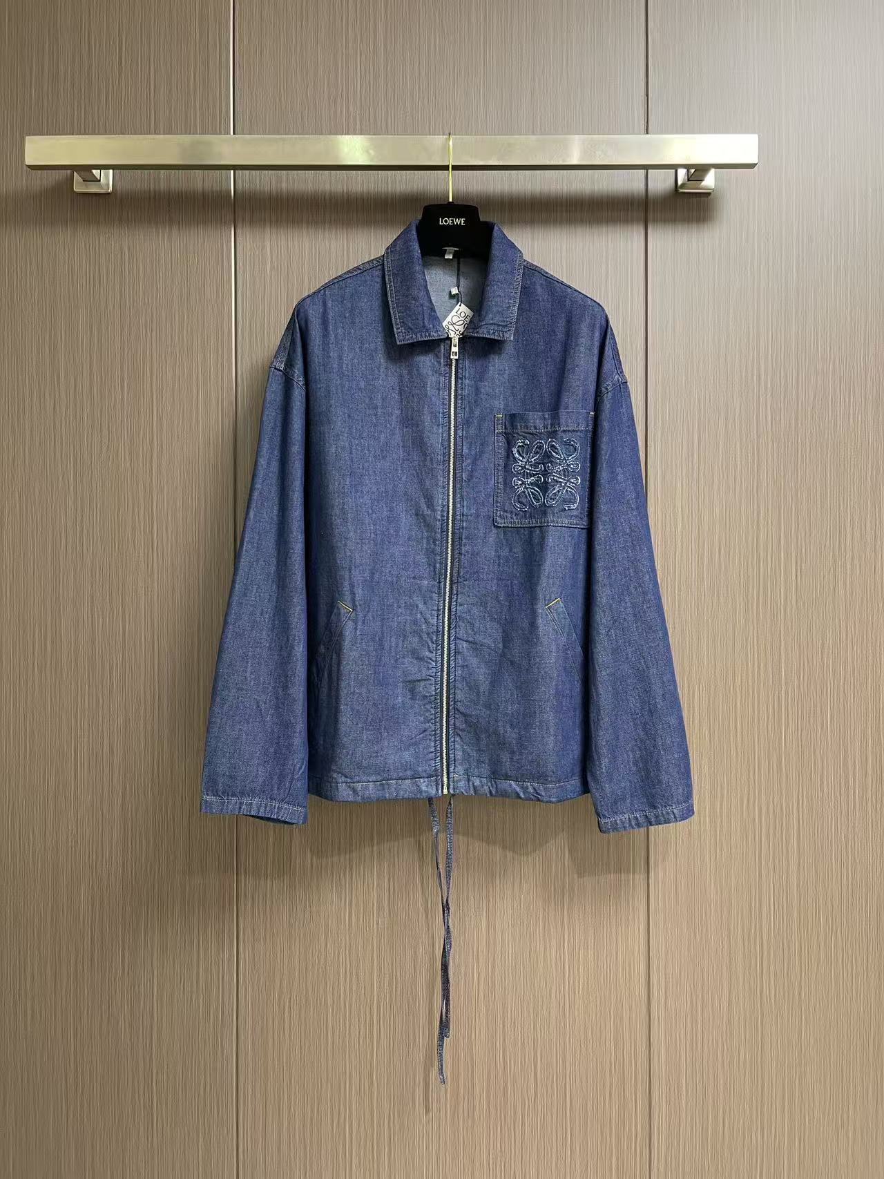 NO:946005,Tops shorts Loewe  Patch pocket embroidered denim jacket and denim shorts set is made of lightweight cotton chambray denim fabric, made with plain weave and close weaving technology. The warp and weft cotton yarns are evenly interwoven, and the fabric surface presents a delicate and clear denim grain texture. It feels soft and thin, and has a moderate drape. It is stiff and not easy to wrinkle and deform. The gray fabric has been washed, pre-shrunk and softened to remove the hard pulpy feel of the fabric, making the upper body light and pressure-free, breathable and breathable.  Sweat-free. The Anagram pattern is created using exclusive appliqué technology. First, the cut-out denim pattern pieces are accurately positioned on the patch bag base, and fixed on the positioning frame of the embroidery machine to prevent deviation. The needle is double-layered and densely embroidered to lock the fabric piece along the outer contour of the pattern, and the special raw edge embroidery stitch method is simultaneously used to outline the edge. The embroidery threads are staggered and stacked to create a fluffy three-dimensional fringe texture, and the multi-layer lock embroidery structure  The patch is firmly fixed, and it is not easy to warp and fall off. The tassel texture is full and three-dimensional. The lines are clean and neat after hand-finishing. It has both three-dimensional visual texture and solid firmness. Classic lapel structure, double-layer panel seaming reinforcement at the collar, double topstitch lock stitching on the inside of the collar, which is not easy to roll up and deform. The embroidered patch pocket on the chest is paired with oblique side pockets on both sides. All pockets are reinforced with double topstitching, and the stitches are tightened at the corners.  The lock knot prevents cracking and deformation when objects are pulled daily, and the diagonal pocket fits the body, taking into account both beauty and practicality. The hem has a built-in cotton drawstring of the same material, which is fixed with metal air eyes, and the tightness of the hem can be adjusted freely; the cuffs are double-layered and hemmed with double sewing threads, and the seams are dense and washable, so there will be no problems with thread breakage or fabric tearing. Sizes S-XL, loewe, shorts, jackets  & coats,19860909上衣 短裤 Loewe 贴袋绣花牛仔外套牛仔短裤套装,选用轻盈棉质青年布牛仔面料,采用平纹紧密梭织工艺打造,经纬棉纱均匀交织,布面呈现细腻清晰的牛仔颗粒肌理,触感柔软轻薄,同时其自带适度垂坠感,挺括不易起皱变形.坯布经水洗预缩软化处理,去除布料硬性浆感,上身轻盈无压,透气排汗不闷肤.采用专属贴布绣工艺打造Anagram纹样,先将裁剪成型的牛仔布图案裁片精准对位铺置在贴袋基底上,固定于绣花机定位框防止走位偏移.针头沿图案外轮廓双层密绣锁固布片,同步运用特殊毛边绣针法勾勒边缘,绣线交错堆叠塑造蓬松立体的流苏肌理,多层锁绣结构牢牢固定贴布,不易翘边脱片,流苏纹理层次饱满立体,手工精修后线条干净利落,兼具立体视觉质感与扎实的牢固度.经典翻领结构,领部双层裁片锁边加固,领口内侧双道明线锁缝,不易翻卷变形.胸前绣花贴袋搭配两侧斜插侧袋,所有袋口全部采用双道明线加固,转角位置收针锁结,日常放置物件拉扯不易开裂变形,斜插袋贴合衣身,兼顾美观与实用性.衣摆内置棉质同料抽绳,搭配金属气眼固定,可自由调节下摆松紧；袖口双层折边双车线缝制,锁边密实耐洗,不会出现断线、布料撕裂问题.尺码S-XL,,loewe,shorts,jackets & coats,,Men's clothing