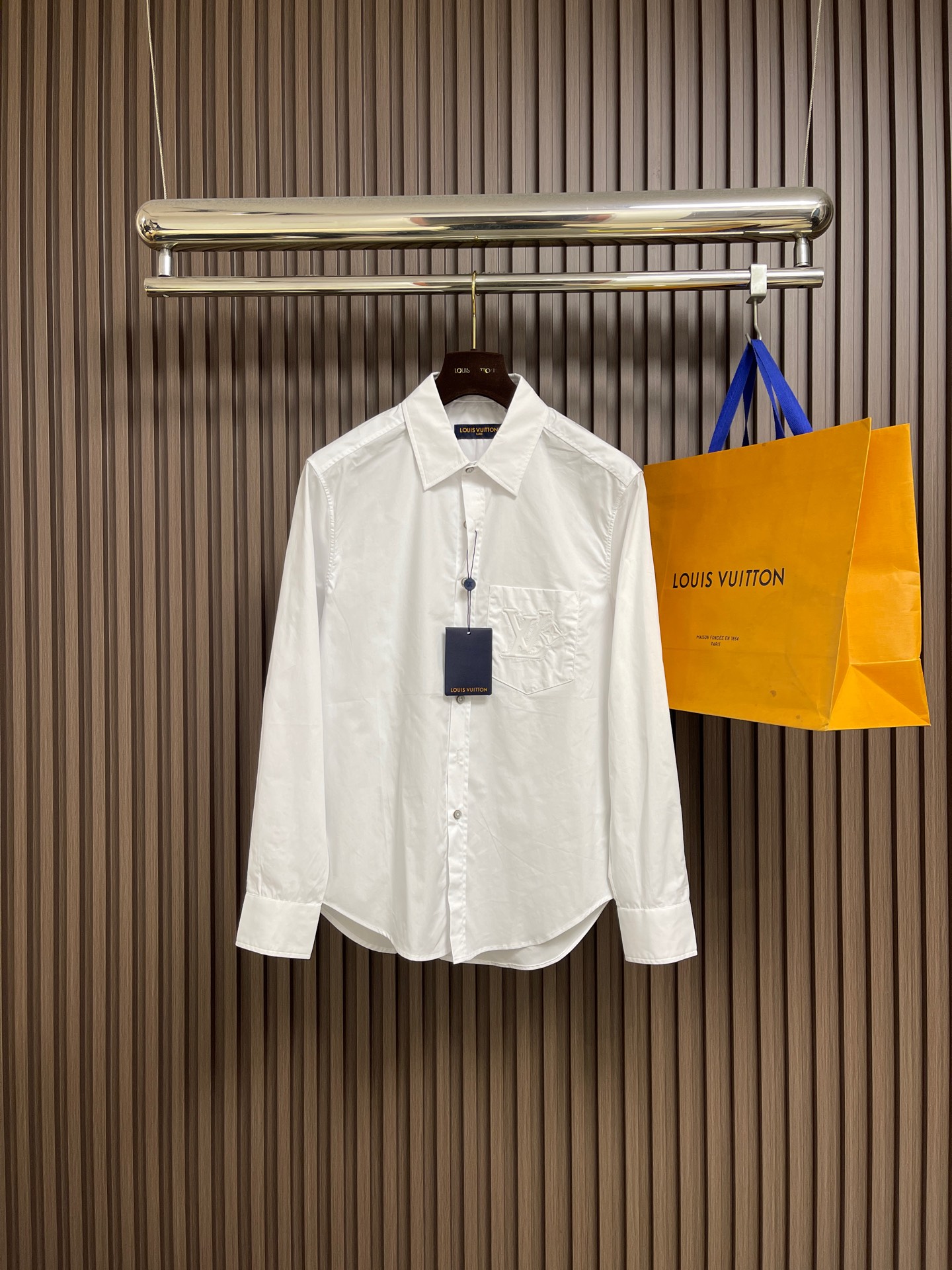 NO:609513,LV  Embossed pocket lapel long-sleeved shirt, twill woven fabric, standard version of peak lapel, hidden front placket, pearlescent buttons at the top, equipped with base, chest pocket with logo embossing, obvious twill texture on the front of the fabric, blurred on the back. Single yarn is used in both the warp and weft directions, the thread density is similar, the warp density of the fabric is slightly higher than the weft density, and the hand feel is relatively soft  Soft. The fabric has excellent stability, is not easy to deform, and prevents bulging; cotton fiber has good hygroscopicity, which keeps the fabric in a water balance state, making people feel comfortable and adding to the sophistication of the fabric. The concave and convex embossing process is heated and shaped by the upper and lower dies, and the rollers press the fabric at a certain temperature, and the production is completed through mechanical embossing.  Produce three-dimensional hierarchical convex surface. The fabric produces a concave and convex pattern with a relief-style three-dimensional effect and a special gloss effect. The three-dimensional shape, pattern and text have a strong three-dimensional effect, adding a decorative art effect, with clear layers, concentrated three-dimensional image, and accurate perspective angle. The collar can be adjusted with buttons for tightness.  Formal feeling and comfort; the rear pleats and ribbons are classic shirting techniques. The pleats provide extra room for movement, and the ribbons can maintain the shape of the shirt when hanging. The button front adopts custom buttons with matching positioning. These details together ensure the practical function and crisp shape of the shirt. Sizes S-XL, Louis  vuitton,shirts,alexander wang19860909LV 压花口袋翻领长袖衬衫,斜纹梭织面料、翻尖领标准版型、隐藏式前襟、顶端珠光纽扣、配有底座、胸袋呈现标识压纹、织物正面斜纹纹路明显,反面比较模糊.经纬向均用单纱,线密度接近,织物经密略高于纬密,手感比较柔软.面料具有极好的稳定性,不易变形、防鼓包；棉纤维具有较好的吸湿性,使织物保持水平衡状态,使人感觉舒适,增添了面料的精致度.凹凸压花工艺由上下模加热定型,轧辊在一定温度下压轧织物,通过机械压印来完成产生立体层次凸面.织物产生具有浮雕风格的立体效应和特别的光泽效果的凹凸花纹的工艺,制作立体造型图案文字有较强的立体感,增添装饰艺术效果,层次清晰、立体形象浓缩、透视角度准确.衣领可通过纽扣调节松紧,兼顾正式感与舒适度；后部褶间与祥带是经典衬衫工艺,褶间提供额外的活动余量,祥带则可在悬挂时保持衬衫形态.纽扣前襟选用定位相符的定制纽扣,这些细节共同确保了衬衫的实用功能与挺括造型.尺码S-XL,,louis vuitton,shirts,alexander wang,Men's clothing