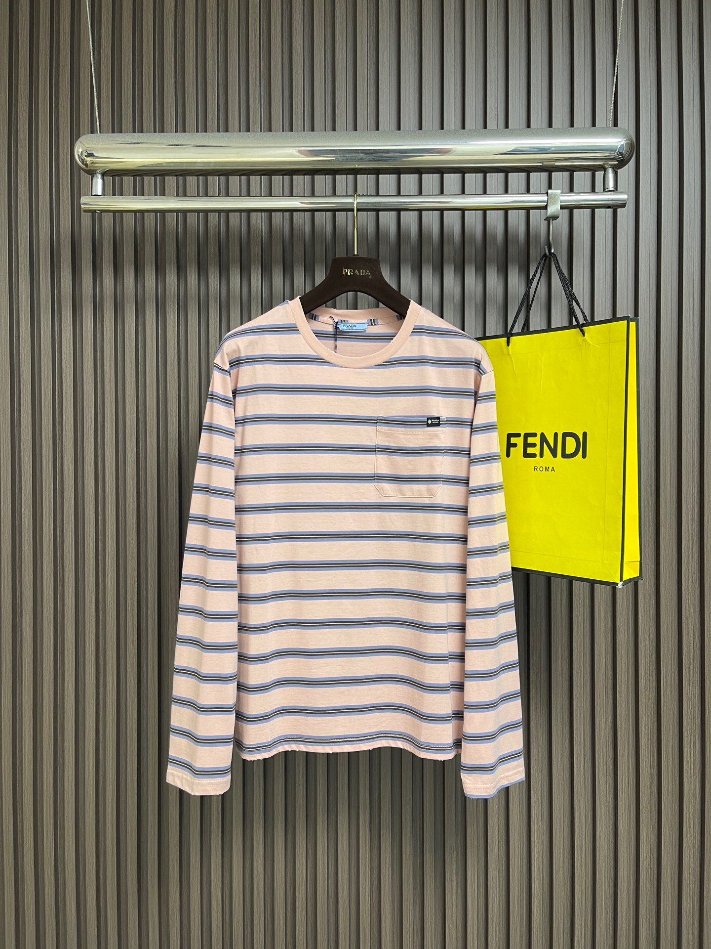 NO:935691,Prada  Pink striped round neck long-sleeved T-shirt, the fabric shows the ultimate lightness in craftsmanship and texture. The compact spinning technology eliminates yarn hairiness through airflow concentration, making the yarn body smooth and round, and the surface of the fabric presents a delicate and high-end texture. The yarn-dying process is the core of this striped T-shirt; the yarn has been pre-dyed as required before weaving.  Blue, white and other colors are then interwoven into striped patterns through a knitting machine. This process is different from weaving first and then dyeing. The colored yarns are interwoven with each other during the weaving process. The stripe boundaries are clear and distinct, and the color fastness is higher. It is not easy to fade or smudge after washing. The 1×1 rib structure gives the fabric extremely high lateral elongation and resilience.  It can be stretched to more than 1.5 times its original length, and can be completely restored to its original shape after release. Positioning and sewing of the entire piece is the most technically difficult process of this T-shirt. The placement of each piece must be strictly aligned with the stripe pattern on the fabric to ensure that the stripes of the entire piece of clothing are continuous and complete after stitching. Cutting  All pieces must be cut in a single layer to ensure that the stripe position of each piece is accurate. When sewing, the stripes at side seams, shoulder seams, armholes and other joints must be completely aligned, which requires the sewing worker to make precise positioning and adjustments during sewing. The overall routing is straight and delicate, the fabric is comfortable and soft, and the colors are consistent without color difference.  Can be matched with the same style to create a fashionable look, to suit various occasions, customized accessories, complete with three standards!  Size XS-S-M-L,,prada,tees,t-shirt,19860909Prada 粉间条纹圆领长袖T恤,面料在工艺与质感上呈现出极致的轻盈感,紧密纺技术通过气流集聚消除纱线毛羽,使纱体光洁圆润,面料表面呈现细腻的高级质感.先染纱工艺是这款条纹T恤的核心；纱线在织造前已预先染成所需的蓝、白等颜色,再通过针织机交织成条纹图案.这种工艺与先织后染不同,色纱在织造过程中相互交织,条纹边界清晰分明,色彩牢度更高,水洗后不易褪色或晕染.1×1罗纹结构赋予了面料极高的横向延伸率与回弹力,横向可拉伸至原长的1.5倍以上,释放后能完全恢复原状.全件定位对线缝制是该款T恤最具技术难度的工艺环节,每一片裁片的摆放位置都必须严格对齐面料上的条纹图案,确保缝合后整件衣服的条纹是连续、完整的.裁剪时所有裁片必须单层裁剪,以保证每一片裁片的条纹位置精确无误.缝制时,侧缝、肩缝、袖窿等拼接处的条纹必须完全对齐,这要求缝纫工在车缝时进行精确的定位与调整.整体走线平直细腻,面料舒适柔软,颜色一致无色差 能与同款搭配营造时尚造型,驾驭各种场合,定制辅料,三标齐全！尺码XS-S-M-L,,prada,tees，t-shirt,,Men's clothing