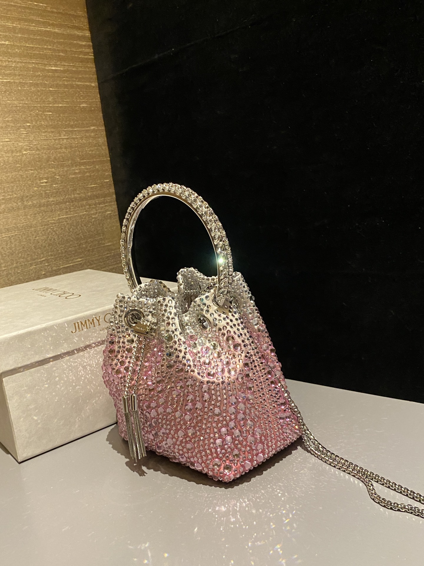 JIMMY CHOO
BON BON系列标志款手提包，光滑缎面铺嵌渐变水晶制作。
设计灵感源于软硬质感及甜美与张扬气质的强烈对比，点睛配以标志性金属流苏，成就如BON BON之名般个性别致又优雅迷人的时尚包袋。现代感桶包廓形，配以手环造型顶部提手，提手饰有百粒经三天精工颗颗细嵌的水晶，精美雅致，招牌奢华包袋当属本款BON BON手提包。
缎面烫钻水晶手嵌水晶饰手环提手Jimmy Choo logo压印金属流苏开合链条肩带内层口袋包袋尺寸：16（长）x15（高）x9.5（宽）厘米肩带长度：55厘米提手长度：9厘米 20259 