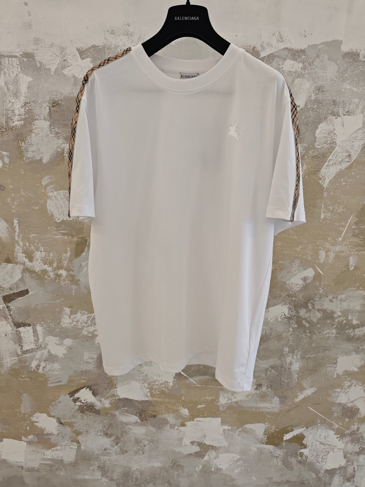 P140 도매 DM 버버리 체크 리본 반팔 티셔츠 색상: 블랙 & 화이트 사이즈: (S, M, XL) 이중 면 평직 원단으로 제작되었습니다. 날실과 씨실이 교차하여 촘촘하면서도 가벼운 균일한 체크 무늬를 만들어냅니다. 면 섬유의 수축 가능성을 적극적으로 제어하여 원단을 안정적인 소재로 변환함으로써 치수 안정성을 향상시키고 변형 및 주름을 줄였습니다. 가슴 부분의 단색 플랫 자수는 고정밀 컴퓨터 자수기를 사용하여 기계 헤드, 바늘대, 스풀의 움직임을 정밀하게 제어합니다. 원단 프레임이 수평 및 수직 방향으로 정확하게 움직여 스티치 궤적을 형성합니다. 위아래 실의 장력을 조절하여 스티치가 평평하고 풀림 없이 깔끔하게 완성되며, ±0.1mm의 오차 범위 내에서 정교한 패턴을 보여줍니다. 1047 Burberry T Shirt