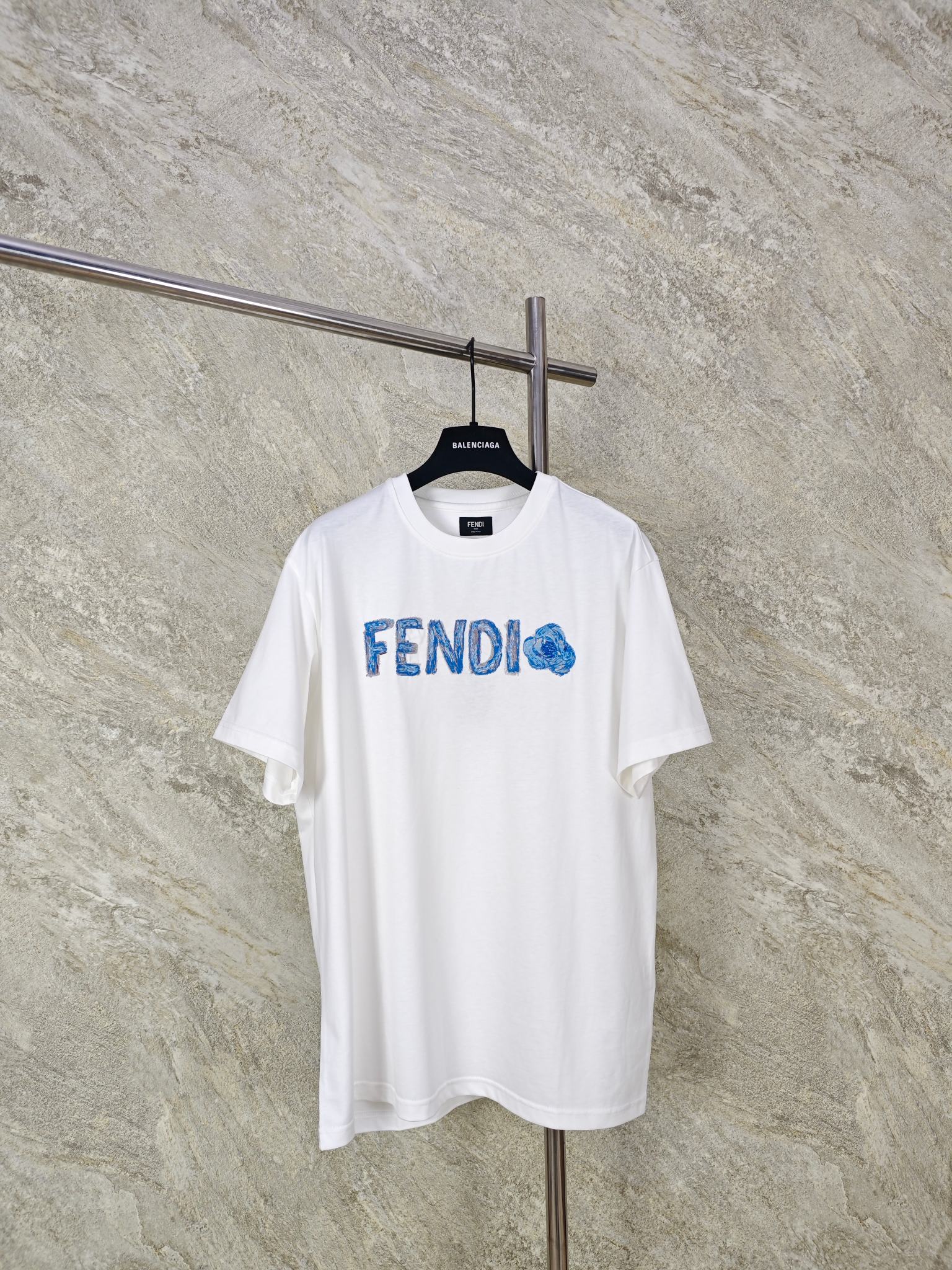 NO:824353,TOP QUALITY FENDI EMBROIDERED SHORT SLEEVE T-SHIRT FABRIC CHOICE  It is made of pure cotton with a weight of 200g. It is derived from natural cotton fiber. It has a soft and delicate touch, is skin-friendly and non-irritating, and can be worn by sensitive skin with peace of mind. It adopts 20S/1 compact spinning process, the yarn has less hairiness and even dryness, and is woven into a plain weave fabric.  The surface is smooth and delicate, the texture is clear and regular, and it is visually more refined and high-end. At the same time, it retains the natural breathable pores of cotton fiber and has excellent moisture absorption and perspiration effects. Compared with ordinary plain cotton, the dense cotton surface is more wear-resistant, less prone to pilling, and more durable for daily wear.  Embroidery technology: The chest is decorated with 21909st's 'FENDI Flower' pattern. It is precision embroidered using a Tajima embroidery machine, integrating multi-color gradients and three-dimensional textures to present a unique watercolor effect. Different shades of blue and white gradient embroidery threads are used for letters and flower patterns, with a natural transition from light blue to indigo, simulating the layering of watercolor smudges. The fineness of each embroidery thread is 40S/2, and high-density embroidery of 1200 stitches/inch ensures uniform color integration.  SizeSMLXL, Fendi-clothing, fendi, fendi, T-shirt, tees, t-shirt,19860909顶级品质 FENDI饰刺绣短袖T恤 面料选择 选用纯棉材质,克重达200g,源自天然棉纤维,触感柔软细腻,亲肤不刺激,敏感肌也能安心穿着.采用20S/1紧密纺工艺,纱线毛羽少、条干均匀,织成的平纹布面光洁细腻,纹理清晰规整,视觉上更显精致高级.同时保留了棉纤维的天然透气孔,吸湿排汗效果出色相比普通平纹棉,紧密棉的布面更耐磨、不易起球,日常穿着更耐造.  绣花工艺 胸前饰以21909st的“FENDI Flower”图案.采用田岛绣花机精密刺绣,融合多色渐变与立体肌理,呈现独特的水彩效果.字母与花朵图案使用不同深浅的蓝白渐变绣线,从浅蓝到靛蓝自然过渡,模拟水彩晕染的层次感.每根绣线细度为40S/2,通过1200针/英寸的高密度刺绣,确保色彩均匀融合 SizeSMLXL,芬迪-服装,fendi,fendi,T-shirt,tees，t-shirt,,Women's clothing