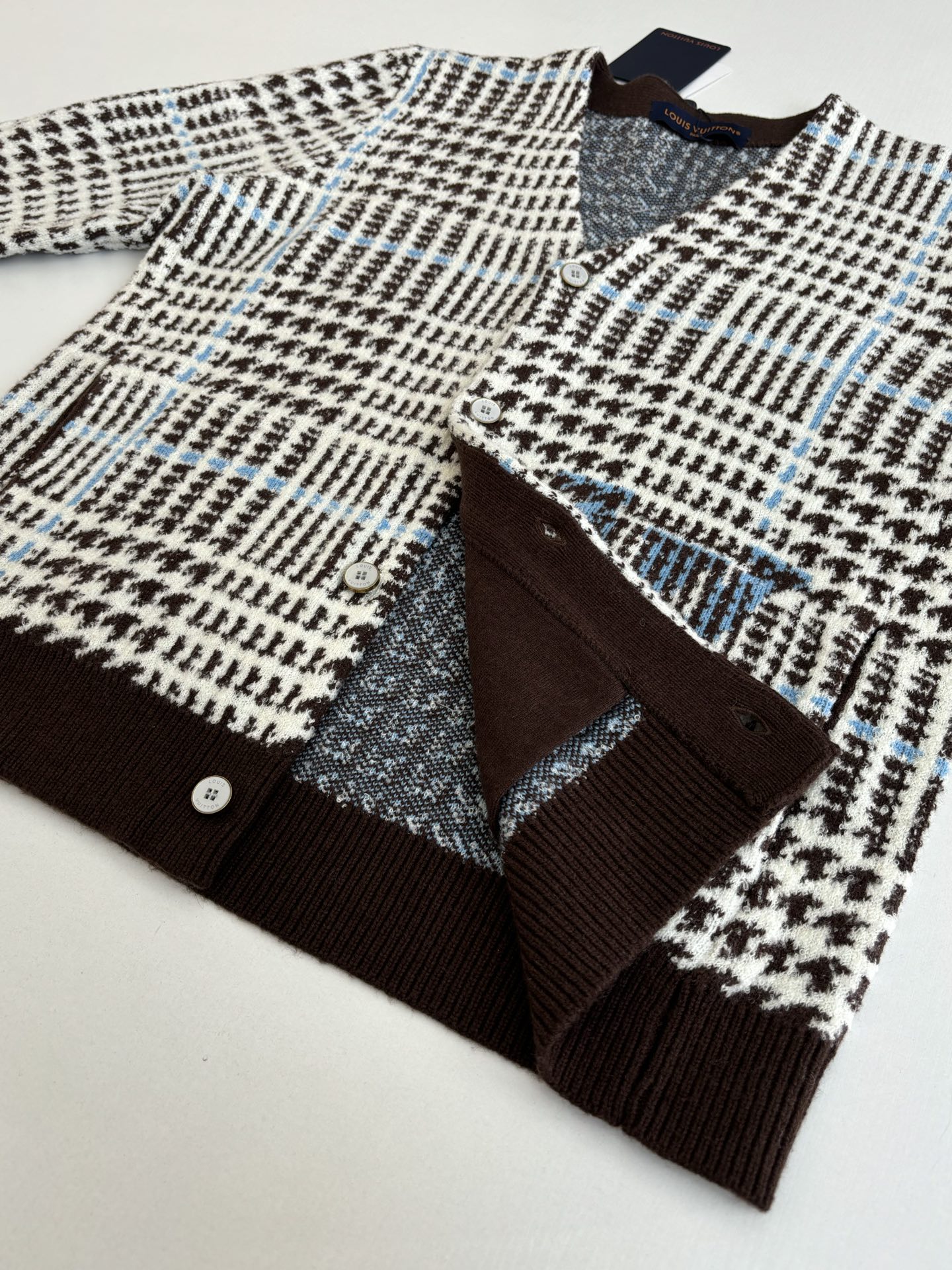 P265
1V/路易 SS26 早春新款 字母嵌花千鸟格针织提花开衫

本款开衫采用全幅提花工艺织造主体图案，以69%绵羊毛混纺31%锦纶的纱线，将千鸟格纹与淡蓝色线条肌理交织呈现，同时通过嵌花工艺在衣身融入品牌标识元素，图案边缘衔接细腻无断层。领口、袖口及下摆选用100%绵羊毛罗纹面料，采用双针锁边工艺收边，兼具弹性与定型效果。前襟配备天然质感纽扣，侧缝口袋采用隐形车线工艺，与衣身提花纹理自然融合。整体版型通过立体剪裁定型，肩部采用无痕缝合工艺，确保穿着贴合度与舒适度，全件由意大利工坊手工细节收尾，呈现高质感针织单品的工艺精度。整体走线平直细腻，面料舒适柔软，颜色一致无色差 能与同款搭配营造时尚造型，驾驭各种场合，定制辅料，三标齐全，男女同款！

Size：S/M/L/XL
