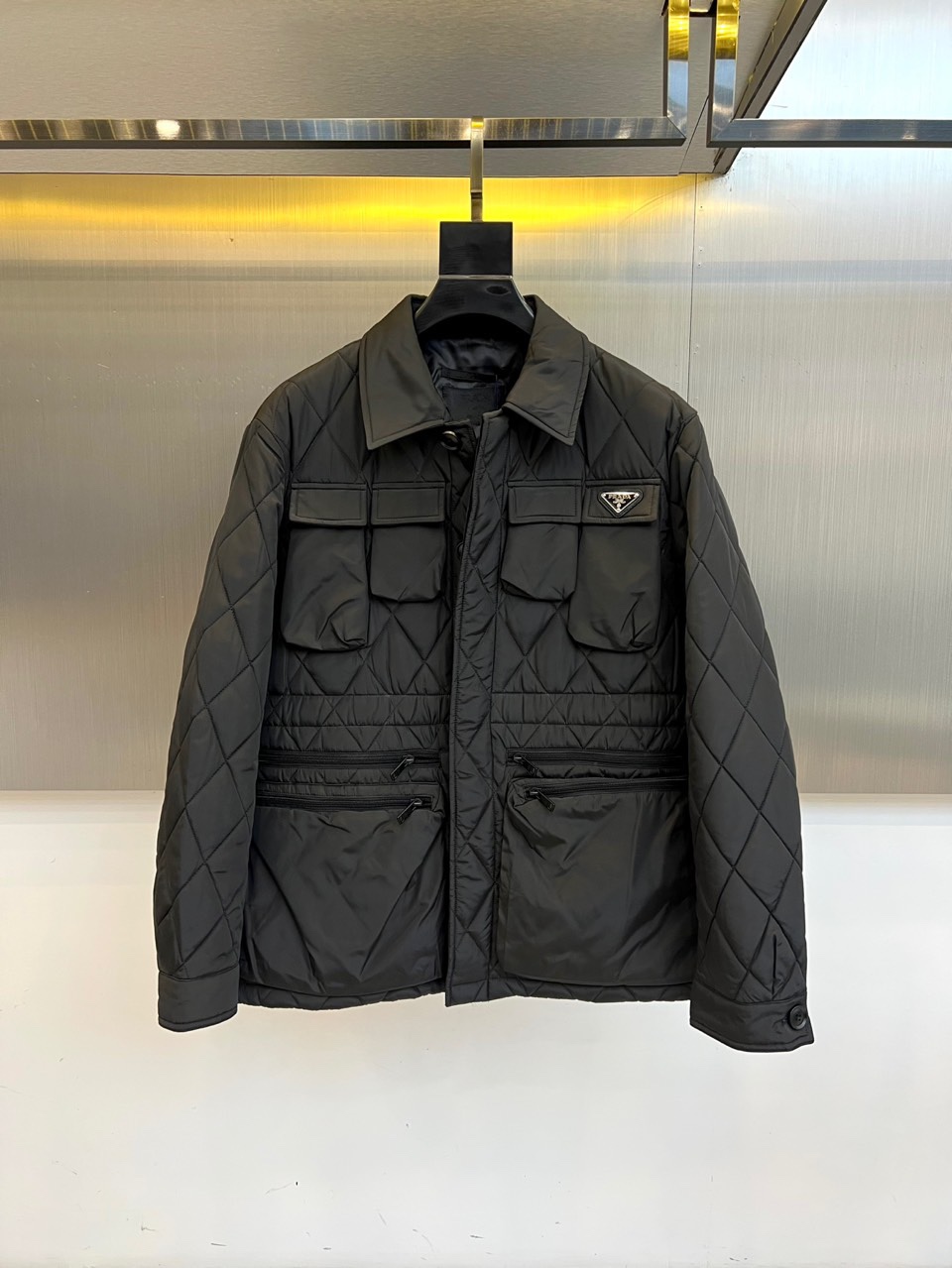 NO:514451,Prad, 25FW autumn and winter new black diamond lapel cotton jacket. The whole body black diamond quilt is like a precision circuit board. The metal triangle logo is scratched on the left chest and is silent. This is the identity chip left by PRADA for the urban dark night. The symmetrical flip pocket and military-grade zipper form a functional array, flowing liquid luster under the warm yellow halo of the metal wall. When the lapel is raised, the neat silhouette reconstructs the body contour. The double zipper pocket can safely store urban survival equipment. Each 0.8cm wide diamond zipper is calculating the optimal light refraction angle.  High-density nylon locks body temperature with architecturally quilted structure. Italian native labels are like secret medals on the inside of the neckline. Hanging in front of the metal background, the jacket itself is a mobile modern art installation. 48-56 (model 173, 143 catties upper body 50 yards), prada, prada, jackets & coats, alexander wang19860909Prad普拉、25FW秋冬新品黑色菱格纹翻领棉服夹克 通体黑色菱格绗缝如精密电路板,金属三角徽标在左胸划破沉寂——这是PRADA留给都市暗夜者的身份芯片.对称翻盖口袋与军工级拉链构成功能主义阵列,在金属墙面的暖黄光晕下流动液态光泽. 翻领立起时,利落剪影重构身体轮廓 双拉链口袋可安全收纳都市生存装备 每道0.8cm宽的菱格都在计算最佳光线折射角度 高密度尼龙以建筑般的绗缝结构锁住体温,意大利原产标签在领口内侧如隐秘勋章.悬挂于金属背景前,这件夹克本身就是移动的现代艺术装置.48-56（模特173,143斤上身50码）,,prada,prada,jackets & coats,alexander wang,Men's clothing