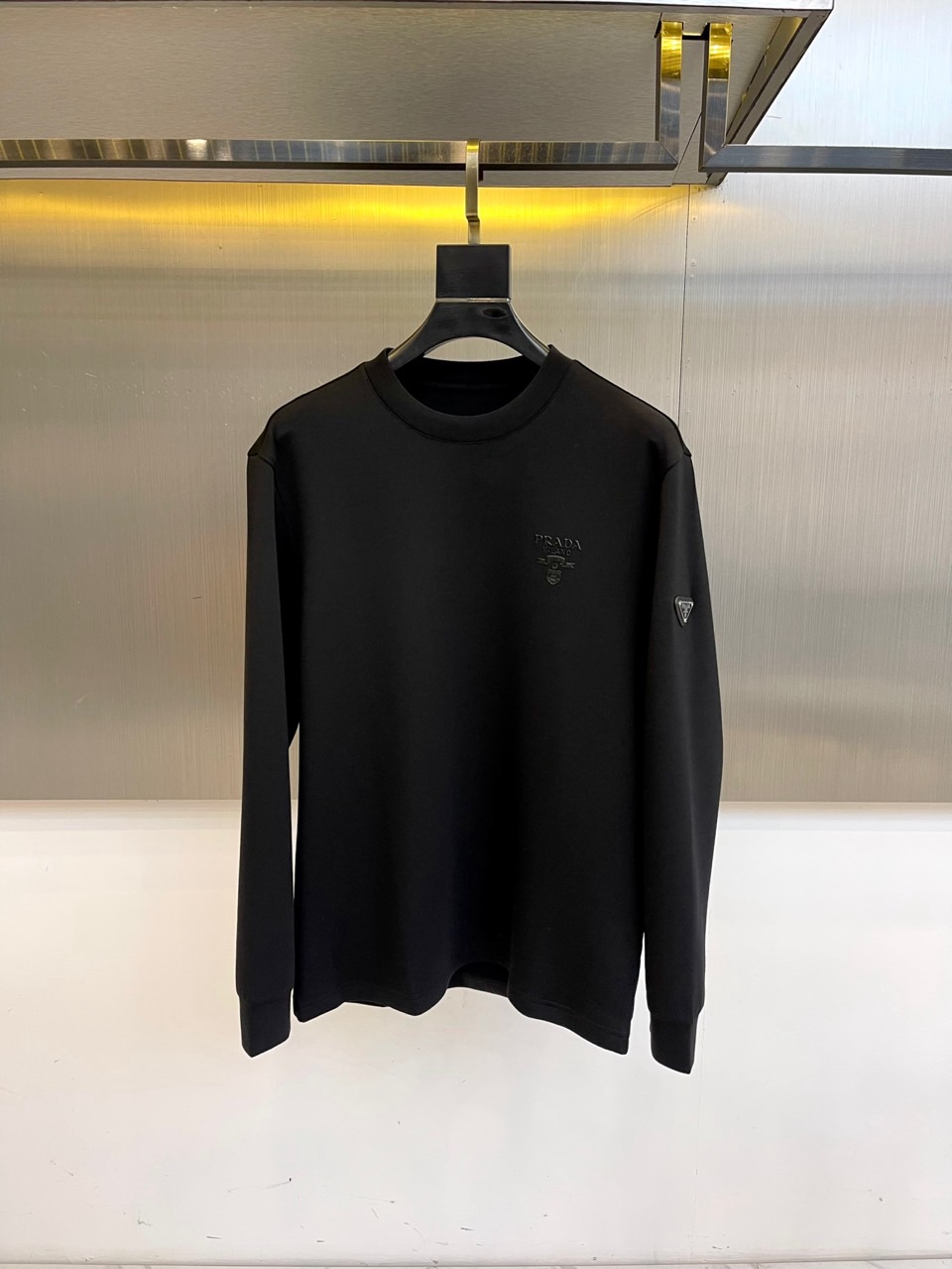 Prad普拉、2025秋冬长袖T恤卫衣。左胸椭圆形金色徽标镌刻品牌基因，领口内侧格纹暗藏经典传承——这是