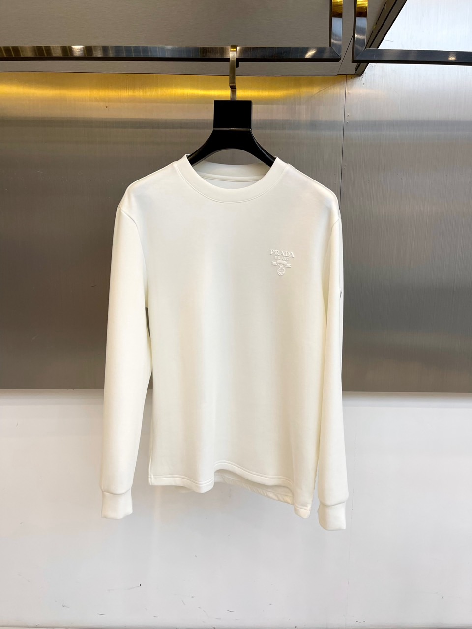 Prad普拉、2025秋冬长袖T恤卫衣。左胸椭圆形金色徽标镌刻品牌基因，领口内侧格纹暗藏经典传承——这是
