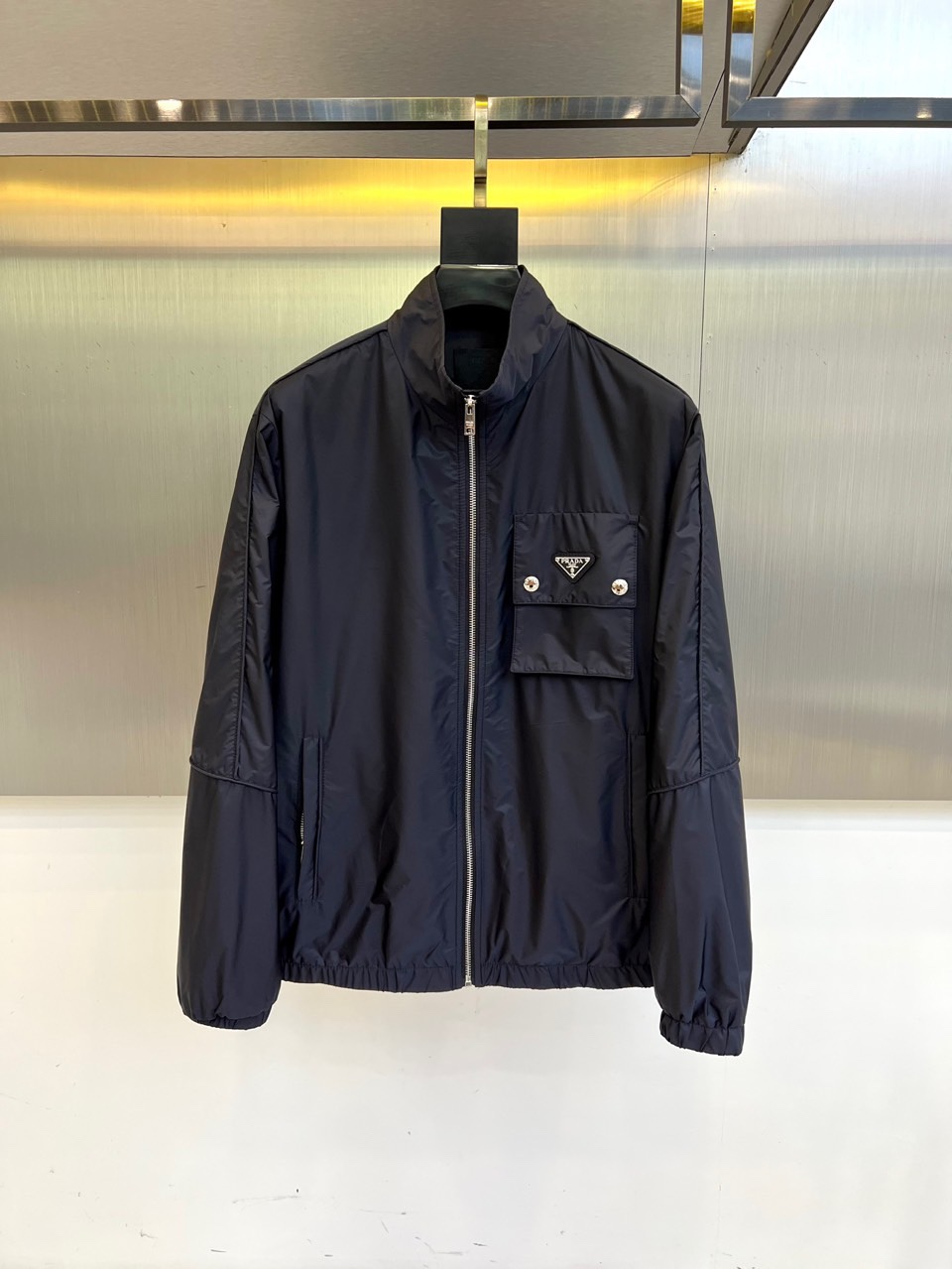 NO:539063,Prad, the new P-home stand-up collar jacket in 2025 autumn and winter, interprets modern minimalist aesthetics in deep navy blue. It uses high-tech nylon fabric to create a light and tough texture. The stand-up collar design is matched with a smooth metal zipper closure, and the left chest is decorated with a brand triangle logo embossed pocket, and two silver buttons embellished details. The cuffs and hem use ribbed closure technology to enhance wind resistance and fit. The tailoring continues Prada's signature neat silhouette, with precise and smooth shoulder lines, and the body is moderately loose and free of movement. The lining is printed  The brand logo is dark, showing low-key luxury. Suitable for urban commuting, business leisure and high-end social occasions, it can be paired with formal pants to create a modern business style, or collided with casual items to create a modern mixed-match aesthetic. It combines practical functions with high-end fashion language with Italian precision techniques, and shows the brand's core concept through detailed design, becoming an iconic item with a sense of technology and luxury in the autumn and winter wardrobe. 48-56 (model 173, 143 catties upper body 50 sizes),, prada, prada, jackets  & coats,alexander wang19860909Prad普拉、2025秋冬新品P家立领夹克,以深邃海军蓝诠释现代极简美学.采用高科技尼龙面料打造轻盈而坚韧的质感,立领设计搭配顺滑金属拉链开合,左胸饰有品牌三角徽标压纹口袋,配以两颗银色纽扣点缀细节.袖口与下摆采用罗纹收口工艺,增强防风性能与贴合度.剪裁延续Prada标志性利落廓形,肩部线条精准流畅,衣身适度宽松兼顾活动自由度.内衬印有品牌标识暗纹,彰显低调奢华.适合都市通勤、商务休闲与高端社交场合,可搭配正装裤塑造摩登商务风范,或与休闲单品碰撞出现代混搭美学.以意大利精工技艺将实用功能与高级时装语言相融合,通过细节设计展现品牌核心理念,成为秋冬衣橱中兼具科技感与奢华质的标志性单品.48-56（模特173,143斤上身50码）,,prada,prada,jackets & coats,alexander wang,Men's clothing