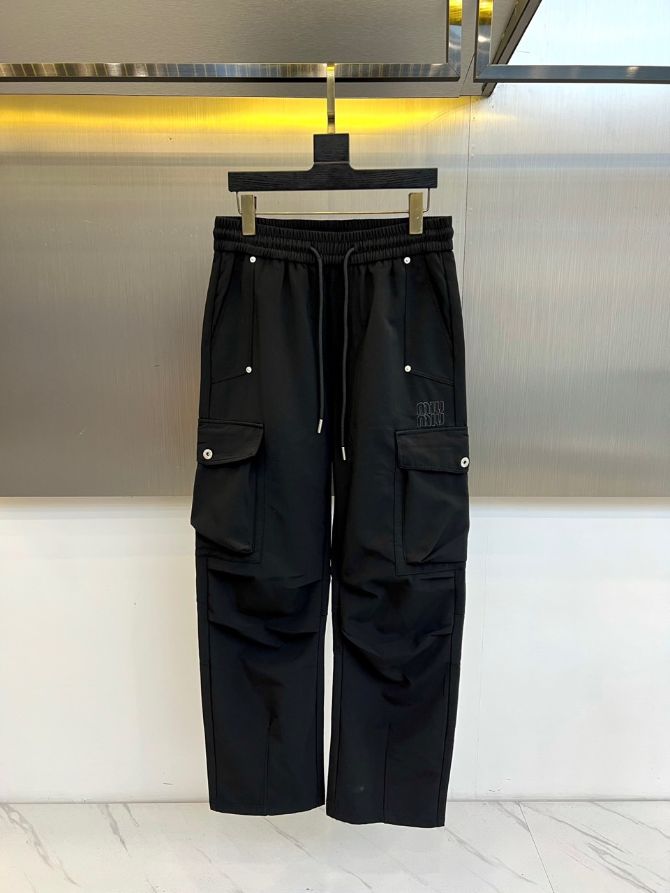 NO:529013,Miu Miu 2025 autumn and winter new black work style trousers are made of the iconic elastic waist and adjustable drawstring design to achieve a personalized fit experience. Both sides of the trousers are equipped with functional large-capacity pockets, and the pockets are decorated with silver rivets, which combine practicality and industrial aesthetic details. The right trousers are embroidered with the brand's iconic letter embroidery, showing the identity with delicate needles. High-density cotton blend fabric is used to present a matte black texture while maintaining an excellent drape. Straight-pin tailoring combined with the design of the trousers legs creates a neat and street-like contour lines. The inside of the waist is made of skin-friendly material to ensure comfort.  The overall style perfectly combines functional style and exquisite aesthetics, and the rivet elements and embroidered logos form a visual tension that combines hardness and softness. The black main tone gives a strong matching possibility, which can be matched with leather short boots to enhance the street attitude, or high heels to create a mixed-match charm. From daily street outing to fashionable parties, it can show unique personality, interpreting Miu Miu's innovative spirit of transforming practical work clothes into high-end fashion. It is recommended to wash and dry cold water to keep the color of the black fabric and the lasting luster of the rivet decoration. 48-56 (model 173, 143 catties upper body 50 sizes),,miumiu,boots,high heels,alexander wang19860909Miu Miu 2025秋冬新品黑色工装风长裤,以标志性松紧腰头搭配可调节抽绳设计,实现个性化贴合体验.裤身两侧配备功能性大容量口袋,袋口饰有银色铆钉装饰,兼具实用性与工业美学细节.右侧裤腿点缀品牌标志性字母刺绣,以细腻针法彰显身份. 采用高密度棉质混纺面料,呈现哑光黑色质感的同时保持出色垂坠感.直筒剪裁结合微收裤脚设计,打造利落且富有街头感的轮廓线条.腰头内部采用亲肤材质处理,确保穿着舒适度. 整体风格完美融合机能风与精致美学,铆钉元素与刺绣标识形成刚柔并济的视觉张力.黑色主调赋予极强的搭配可能性,既可搭配皮质短靴强化街头态度,亦可搭配高跟鞋碰撞出混搭魅力.从日常出街到时尚派对皆能展现独特个性,诠释Miu Miu将实用工装转化为高级时装的革新精神.建议冷水手洗并平铺晾干,以保持黑色面料色泽与铆钉装饰的持久光泽.48-56（模特173,143斤上身50码）,,miumiu,boots,high heels,alexander wang,Men's clothing