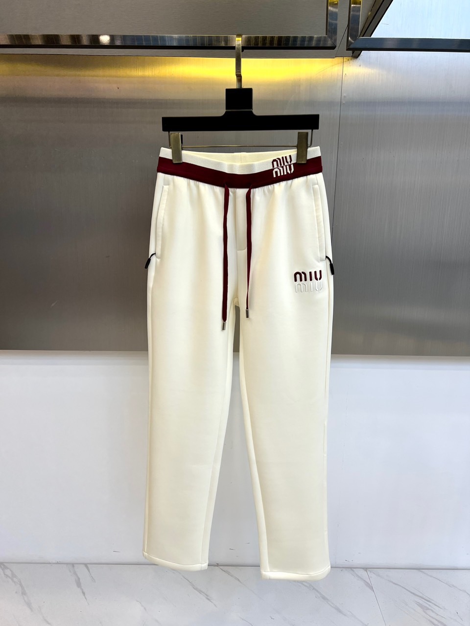 Miu Miu 2025秋冬新品米白色休闲裤，以纯净色调演绎现代少女式叛逆。腰头处饰有酒红色镶边撞色设计