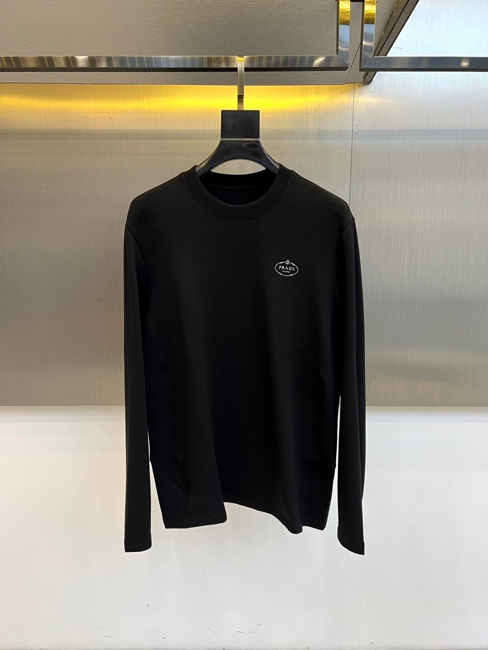 Prad普拉、2025秋冬新品P家简约长袖上衣这款白色长袖上衣采用经典圆领设计与立体剪裁，左胸处装饰品牌