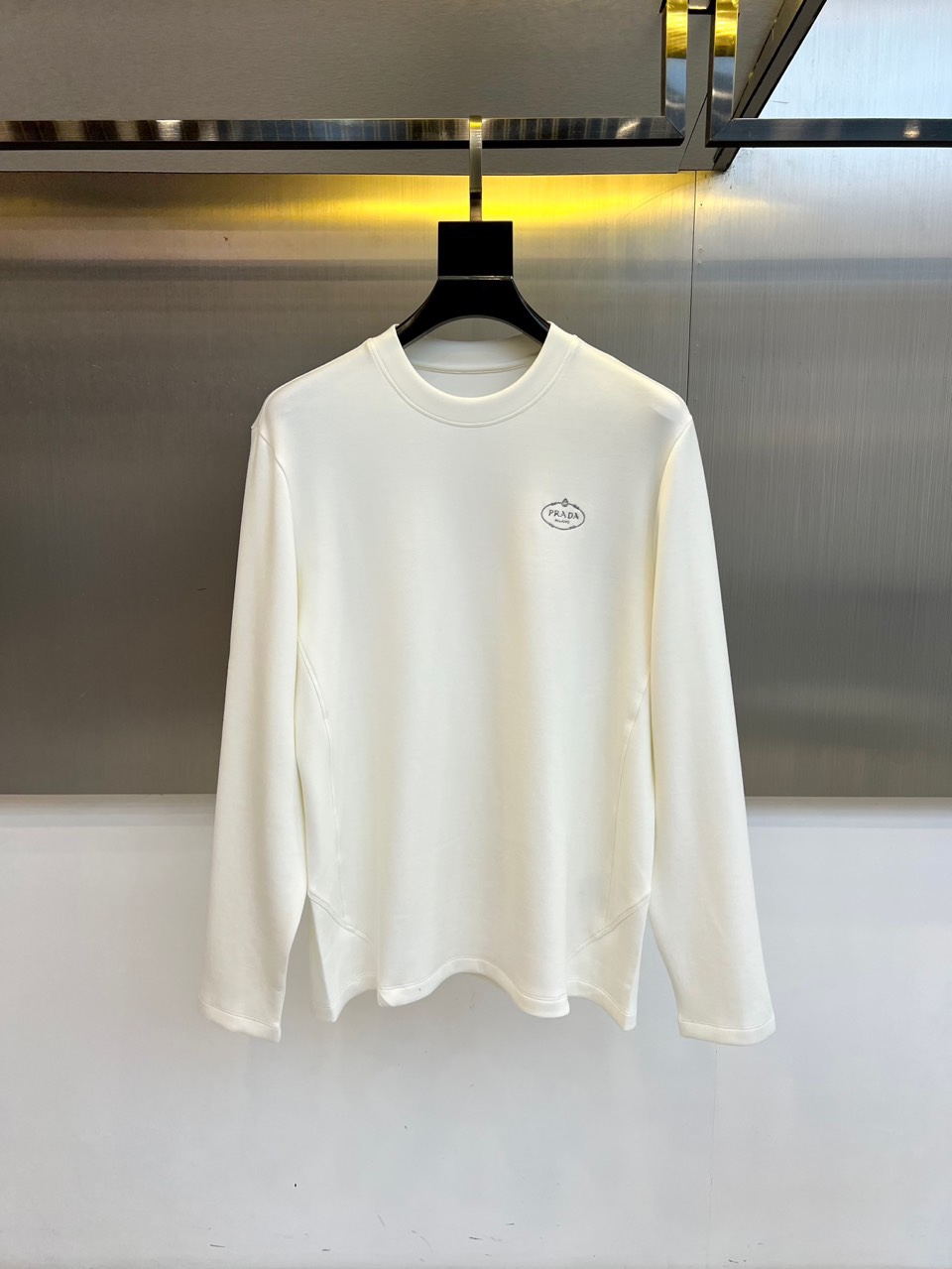 NO:545559,Prad, the new product for autumn and winter, P's simple long-sleeved top. This white long-sleeved top adopts a classic round neck design and three-dimensional tailoring. The brand's iconic oval logo is decorated on the left chest to show low-key luxury. The unique curved tailoring of the hem breaks the conventional design and injects a modern fashion sense into the simple style. The fabric is made of 74.5 viscose fiber, 20.8 nylon and 4.7 spandex in a scientific ratio, which is both soft and skin-friendly and has good elasticity. The ribbed cuffs and collar keep the shape crisp. The curved hem is  The design not only enhances the visual layering, but also enhances the freedom of movement. Whether it is paired with formal trousers to show a business casual style, or paired with casual trousers to create a simple and fashionable sense, it can show PRADA's unique Italian aesthetics through minimalist design language. The fabric has excellent drape, and the white basic model is easy to match, perfect for various occasions. 48-56 (model 173, 143 pounds, upper body size 50),, prada, prada, alexander   wang19860909Prad普拉、秋冬新品P家简约长袖上衣这款白色长袖上衣采用经典圆领设计与立体剪裁,左胸处装饰品牌标志性椭圆形徽标展现低调奢华.下摆独特的弧形剪裁打破常规设计,为简约款式注入现代时尚感.面料选用74.5粘纤、20.8锦纶与4.7氨纶科学配比,兼具柔软亲肤性与良好弹性.罗纹袖口与衣领保持版型挺括.弧形下摆设计不仅提升视觉层次感,更增强活动自由度.无论是搭配正装裤展现商务休闲风,还是搭配休闲裤营造简约时尚感,都能通过极简设计语言展现PRADA特有的意式美学.面料垂坠感优异,白色基础款易于搭配,完美适配多种场合穿着需求.48-56（模特173,143斤上身50码）,,prada,prada,alexander wang,Men's clothing