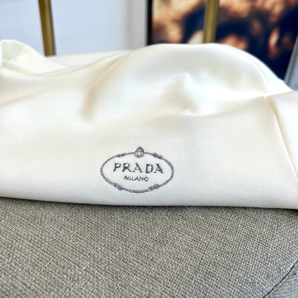 Prad普拉、2025秋冬新品P家简约长袖上衣这款白色长袖上衣采用经典圆领设计与立体剪裁，左胸处装饰品牌