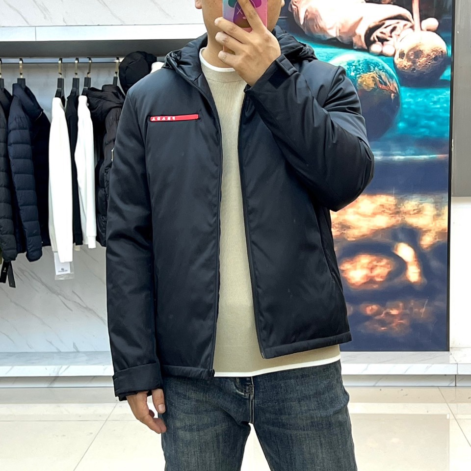Prad普拉、2025秋冬新品 PRADA科技防护羽绒服。深蓝色连帽设计搭配精密拉链系统，左胸红色织带镶