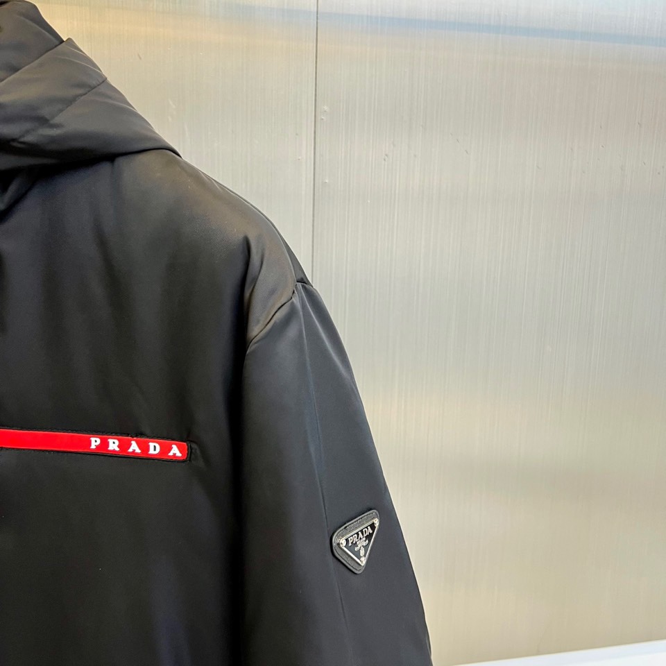 Prad普拉、2025秋冬新品 PRADA科技防护羽绒服。深蓝色连帽设计搭配精密拉链系统，左胸红色织带镶