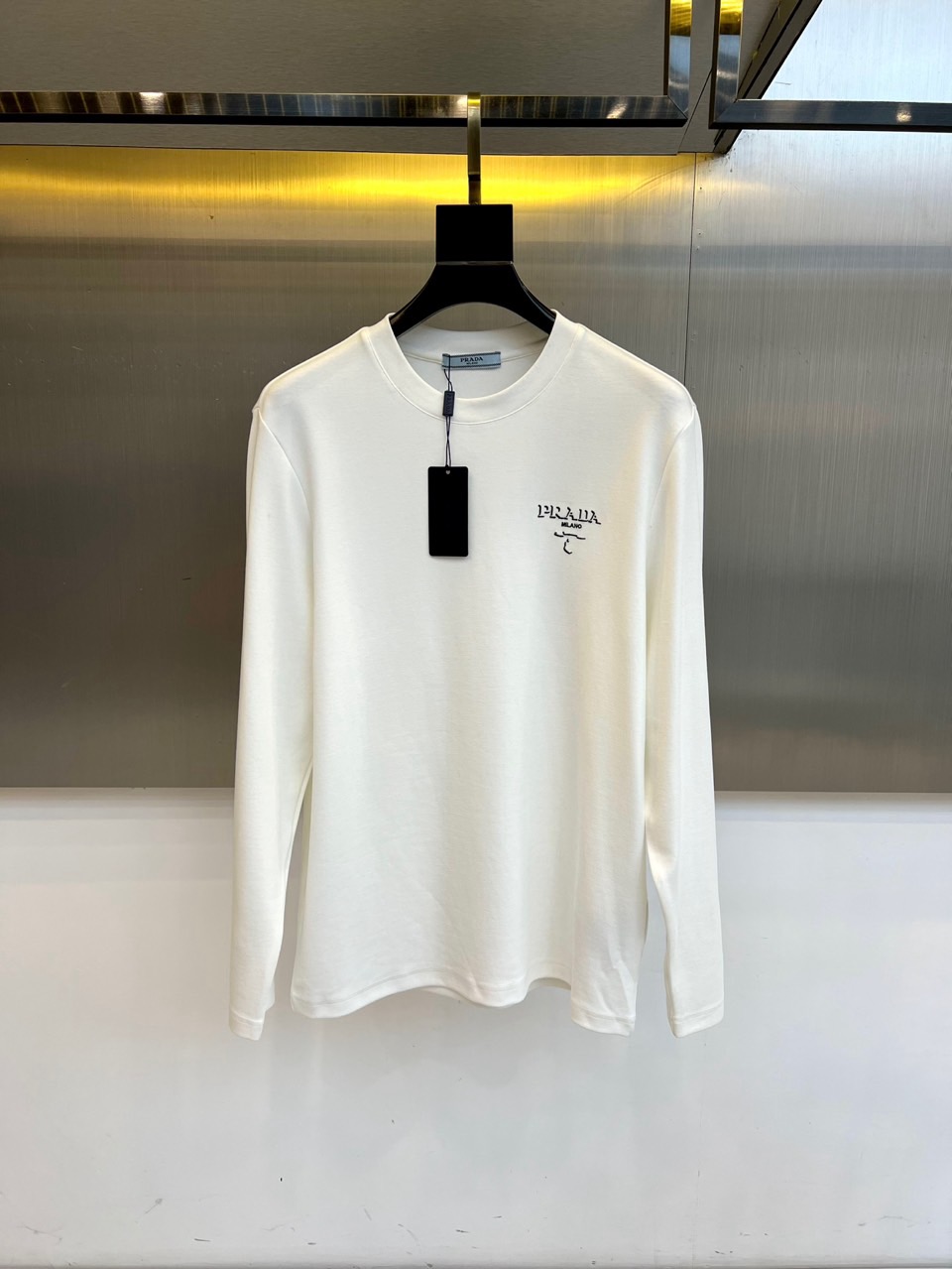 NO:599937,Prada, the 2025 autumn and winter new round-neck long-sleeved top, interprets Spanish luxury aesthetics with minimalist design. The style adopts a classic round-neck silhouette, with the brand's iconic Anagram pattern embroidery on the left chest, and delicate stitching to demonstrate exquisite craftsmanship. The body is made of high-quality cotton material to ensure a skin-friendly and breathable experience, and the ribbing process keeps the shape crisp. Its design integrates traditional leather craft genes into daily wear, and can be paired with casual pants alone.  Showing simplicity and elegance, it can also be layered as an inner layer with a coat to create a sense of hierarchy. From urban commuting to social occasions, this top with pure white tone and iconic details has become a fashion carrier connecting the brand's century-old heritage and modern life, interpreting LOEWE's eternal pursuit of materials and shapes. 48-56 (model 173,143 pounds, upper body size 50),, loewe, prada, alexander  wang19860909普拉达、2025秋冬新品圆领长袖上衣,以极简设计诠释西班牙奢华美学.款式采用经典圆领廓形,左胸点缀品牌标志性Anagram图案刺绣,细腻针脚彰显精湛工艺.衣身选用高品质棉质材质,确保亲肤透气体验,罗纹收边工艺保持版型挺括.其设计将传统皮革工艺基因融入日常着装,可单独搭配休闲裤展现简约优雅,亦可作为内搭与大衣层叠穿搭营造层次感.从都市通勤到社交场合,这款上衣以纯净白色调与标志性细节,成为连接品牌百年传承与现代生活的时尚载体,诠释LOEWE对材质与形制的永恒追求.48-56（模特173,143斤上身50码）,,loewe,prada,alexander wang,Men's clothing