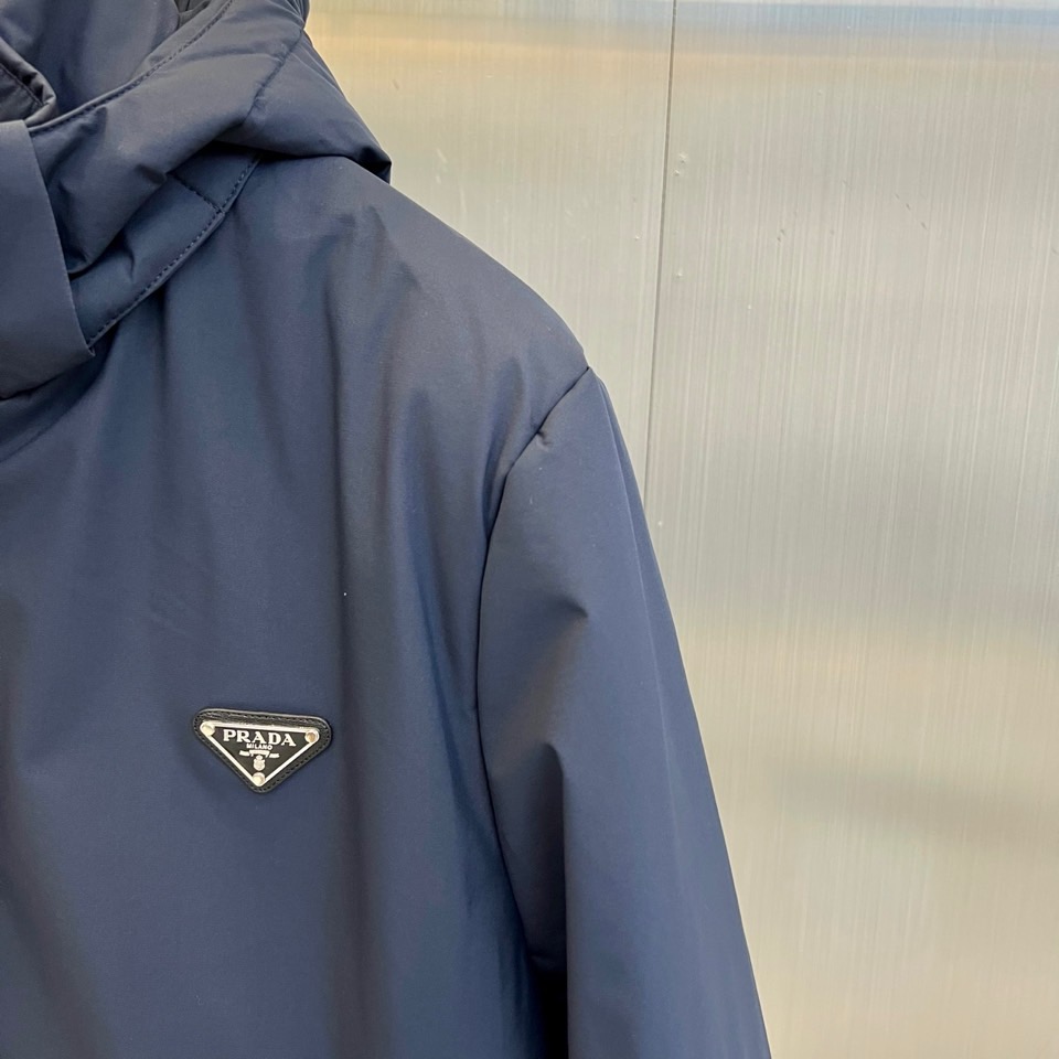 Prad普拉达、2025秋冬新款棉服以深邃藏蓝色演绎都市实用美学。这款连帽棉服采用立体绗缝工艺形成规整的