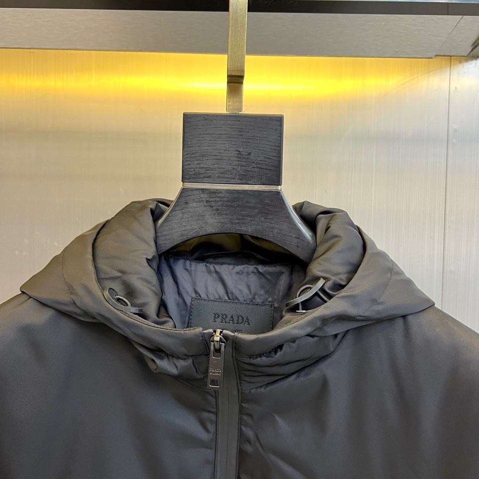 Prad普拉、2025秋冬新品 PRADA科技防护羽绒服。深蓝色连帽设计搭配精密拉链系统，左胸红色织带镶