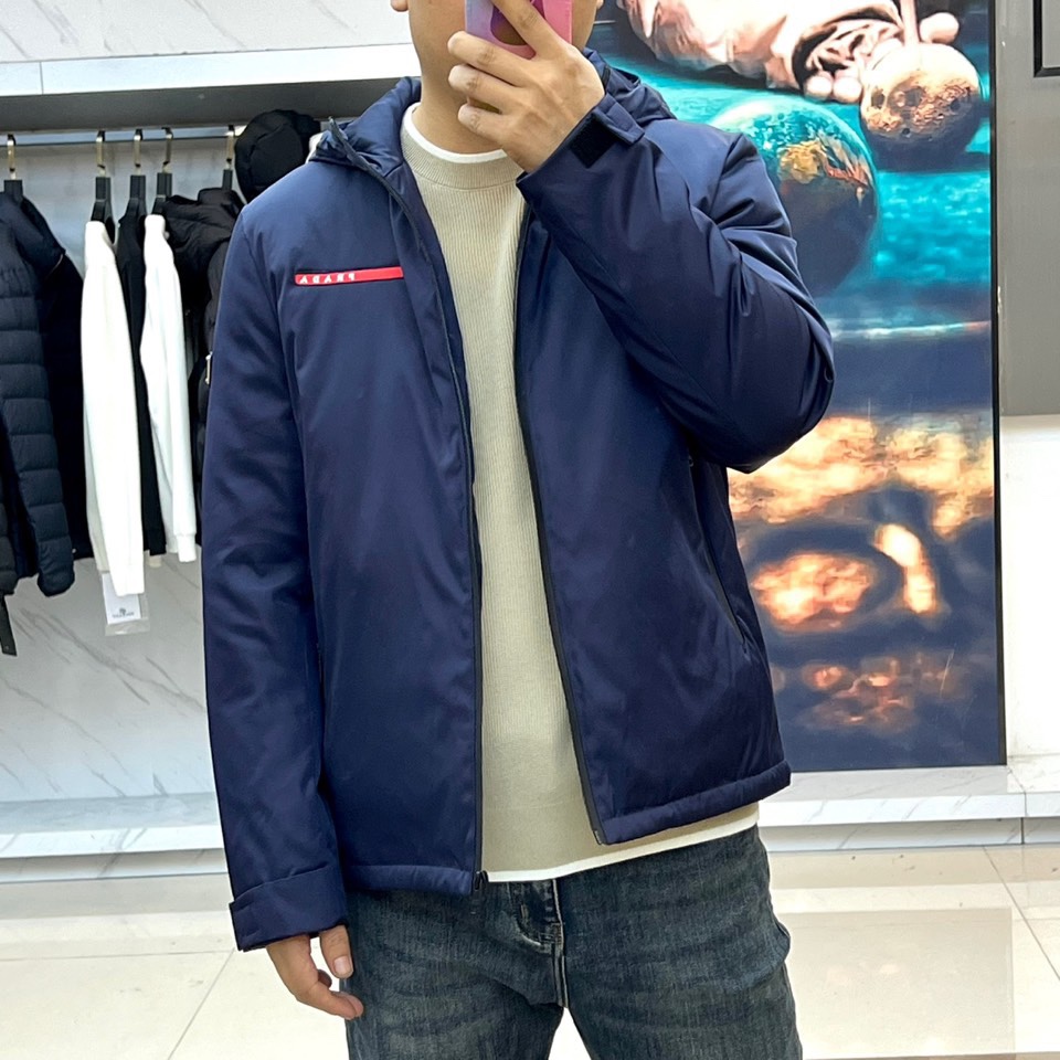 Prad普拉、2025秋冬新品 PRADA科技防护羽绒服。深蓝色连帽设计搭配精密拉链系统，左胸红色织带镶