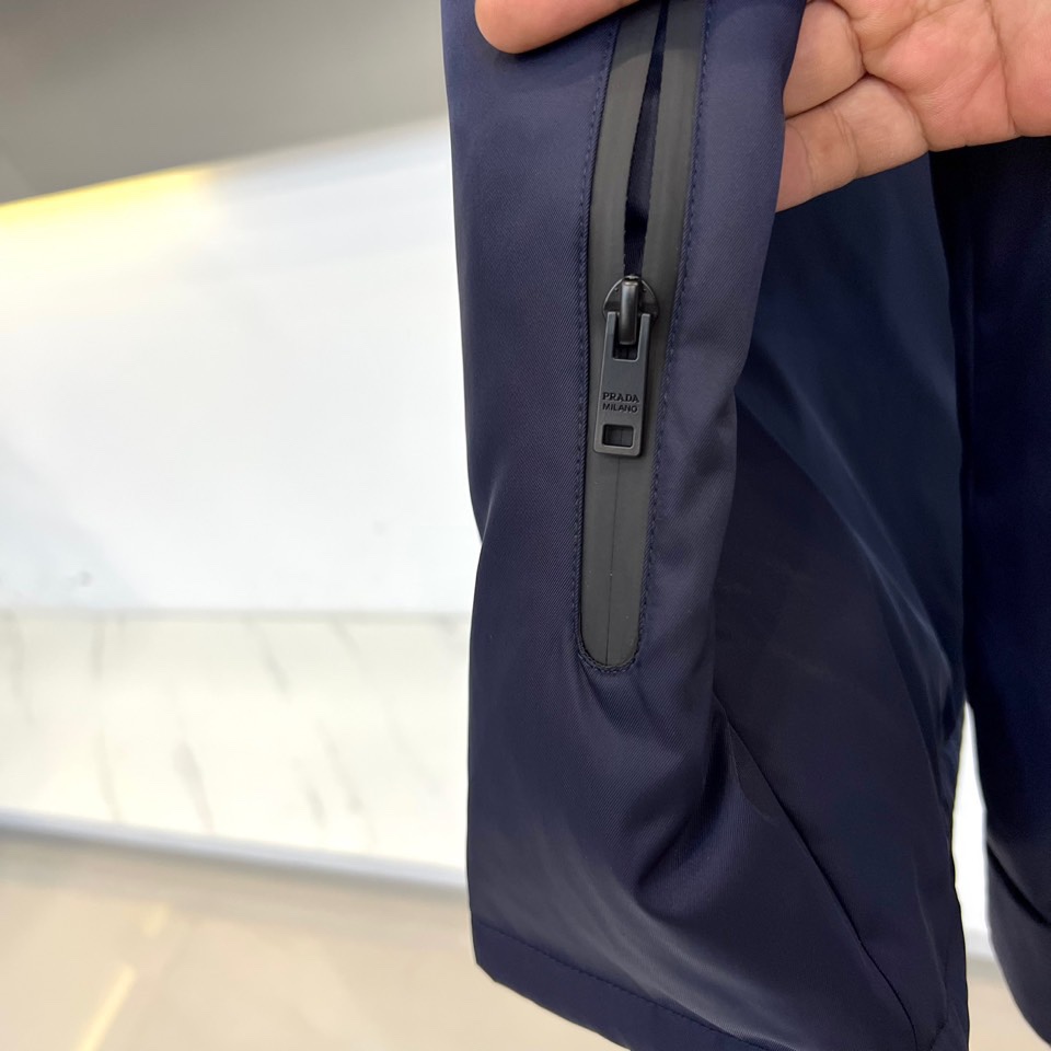 Prad普拉、2025秋冬新品 PRADA科技防护羽绒服。深蓝色连帽设计搭配精密拉链系统，左胸红色织带镶