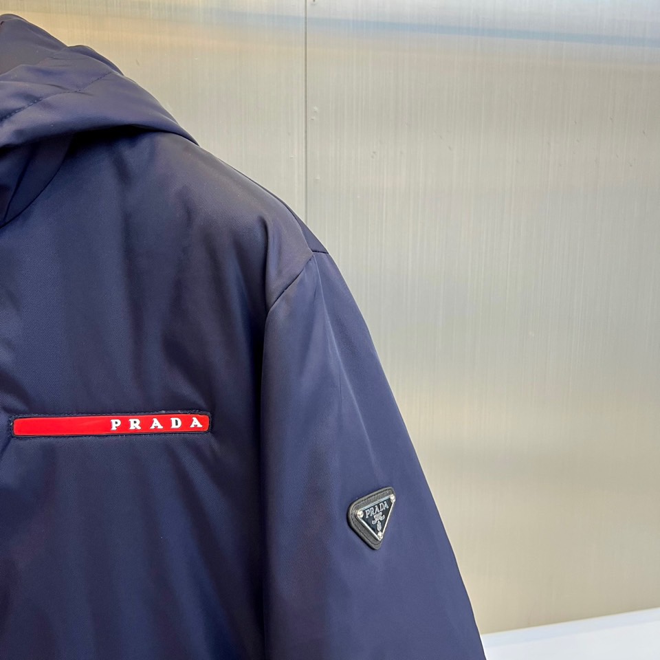 Prad普拉、2025秋冬新品 PRADA科技防护羽绒服。深蓝色连帽设计搭配精密拉链系统，左胸红色织带镶