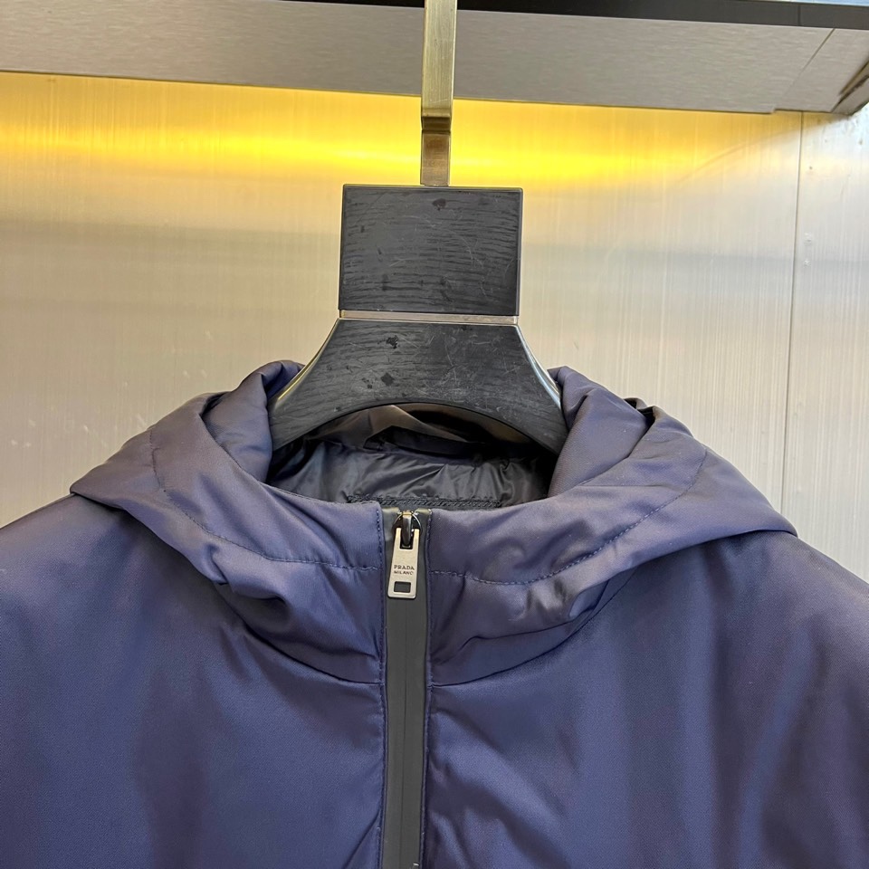 Prad普拉、2025秋冬新品 PRADA科技防护羽绒服。深蓝色连帽设计搭配精密拉链系统，左胸红色织带镶