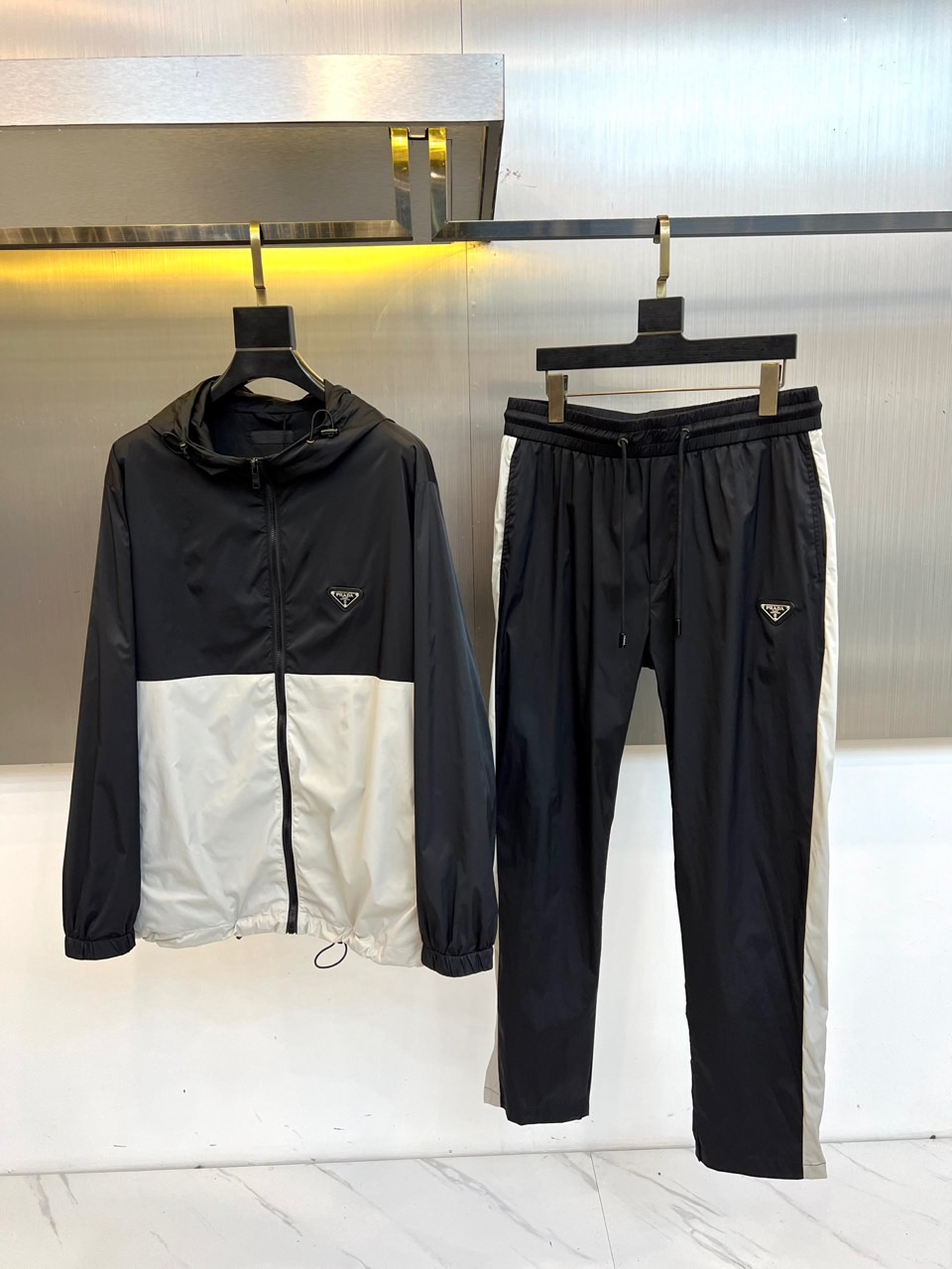 NO:648413,Prad, autumn and winter series of suits. Redefine urban dressing with refined pragmatism language. This sports and leisure suit includes a hooded zipper jacket and a pair of side striped casual trousers. The jacket is spliced with bright black, white and silver technical fabrics to create a three-dimensional vision.  Layered and futuristic silhouette, the left chest is decorated with a classic triangular enamel logo, low-key highlighting the brand identity. The adjustable drawstring hem enhances the functionality of the shape. The trousers are based on black, and the white stripes running through the sides smoothly extend the visual lines, cleverly echoing the color of the top. The drawstring at the waist and left  The details of the micro triangular logo on the legs show ingenuity in simplicity. The suit is made of functional fabrics with subtle luster, and is neatly cut and takes into account dynamic comfort. It combines the precise tailoring of high fashion with the ease of sportswear. The black and white tones are calm and full of power, which perfectly interprets Prada.  Da Shi's modern and intelligent aesthetics can easily show a calm and extraordinary contemporary temperament whether it is dealing with busy daily life or pursuing stylized leisure moments. S-XXL (model 173, 143 pounds, upper body size M),, prada, prada, jackets  & coats,hats,alexander wang19860909Prad普拉达、秋冬系列套装.以精炼的实用主义语言重新定义都市着装.本款运动休闲套装包含一件连帽拉链外套与一条侧纹休闲长裤.外套采用鲜明的黑、白、银三色科技感面料拼接,塑造立体的视觉层次与未来感廓形,左胸饰有经典的三角形珐琅徽标,低调彰显品牌身份.可调节抽绳下摆增强造型功能性.长裤以黑色为基调,侧边贯穿的白色条纹流畅延伸视觉线条,与上衣配色巧妙呼应.腰部抽绳与左腿的微型三角标细节,于简约中见匠心.套装采用具有微妙光泽的功能性面料,剪裁利落且兼顾动态舒适.它将高级时装的精准剪裁与运动服饰的自在感合二为一,黑白色调冷静而充满力量,完美诠释了普拉达式的摩登智能美学,无论是应对繁忙日常或追求风格化的休闲时刻,皆能游刃有余地展现从容不凡的当代气质.S-XXL（模特173,143斤上身M码）,,prada,prada,jackets & coats,hats,alexander wang,Men's clothing