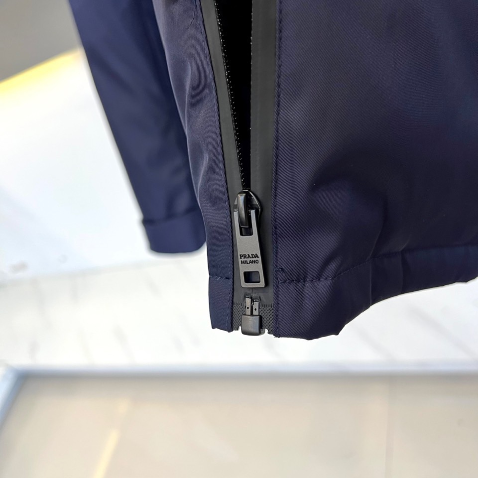 Prad普拉、2025秋冬新品 PRADA科技防护羽绒服。深蓝色连帽设计搭配精密拉链系统，左胸红色织带镶