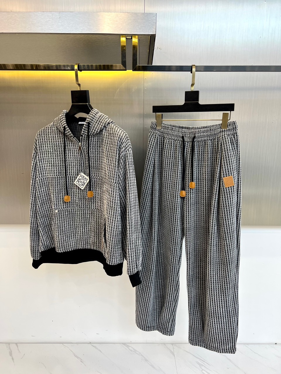 NO:681016,The LOEWE suit features the classic black and white houndstooth pattern throughout, subtly combining retro elegance with modern fashion sense. The houndstooth pattern on the hooded top is delicate and eye-catching, and is cleverly spliced with black fabric from the collar to the cuffs, with a neat outline. There are pockets on the chest, both functional and designed. The most eye-catching thing is the white drawstring and the delicate gold squares at the end, which create unique highlights on the dark plaid, injecting a touch of uninhibited agility into the loose fit.  The matching wide-leg trousers continue the same pattern, with an elastic waistband to ensure comfort, and the same white drawstring and gold square decorations echo the same pattern. The light-colored patch stitching on the side of the trouser legs breaks the continuity of the pattern, using deconstruction techniques to bring unexpected details and interest, showing the brand's low-key and playful personality.  The silhouette of the entire suit is relaxed and moderate, and rich layers are created through material contrast, detailed decoration and structural ingenuity in the classic pattern. Hanging in front of a simple background, the texture and design language of the clothing itself become the absolute focus, highlighting Loewe's unique style of integrating traditional craftsmanship and contemporary aesthetics. M-3XL (model 173-140 pounds upper body size L),,loewe,loewe,hats,19860909罗意威（LOEWE）套装,以经典的黑白千鸟格纹贯穿始终,将复古优雅与现代时尚感精妙结合. 连帽上衣的千鸟格图案细腻醒目,从领口到袖口巧妙拼接黑色面料,轮廓利落.胸前设有口袋,功能与设计兼具.最引人注目的是白色抽绳与末端精巧的金色小方块,在深色格纹上跳跃出别致亮点,为宽松版型注入一丝不羁的灵动. 与之相配的阔腿裤延续了同一纹样,松紧腰头确保了穿着的舒适度,同款白色抽绳与金色方块装饰与之呼应.裤腿一侧的浅色补丁式拼接,打破了图案的连续性,以解构手法带来意想不到的细节趣味,展现了品牌低调玩味的个性. 整套套装廓形松弛有度,在经典图案中通过材质对比、细节装饰与结构巧思营造丰富层次.悬挂于简洁背景前,服装本身的质感与设计语言成为绝对焦点,彰显罗意威融合传统工艺与当代美学的独特风格.M-3XL（模特173-140斤上身L码）,,loewe,loewe,hats,,Men's clothing