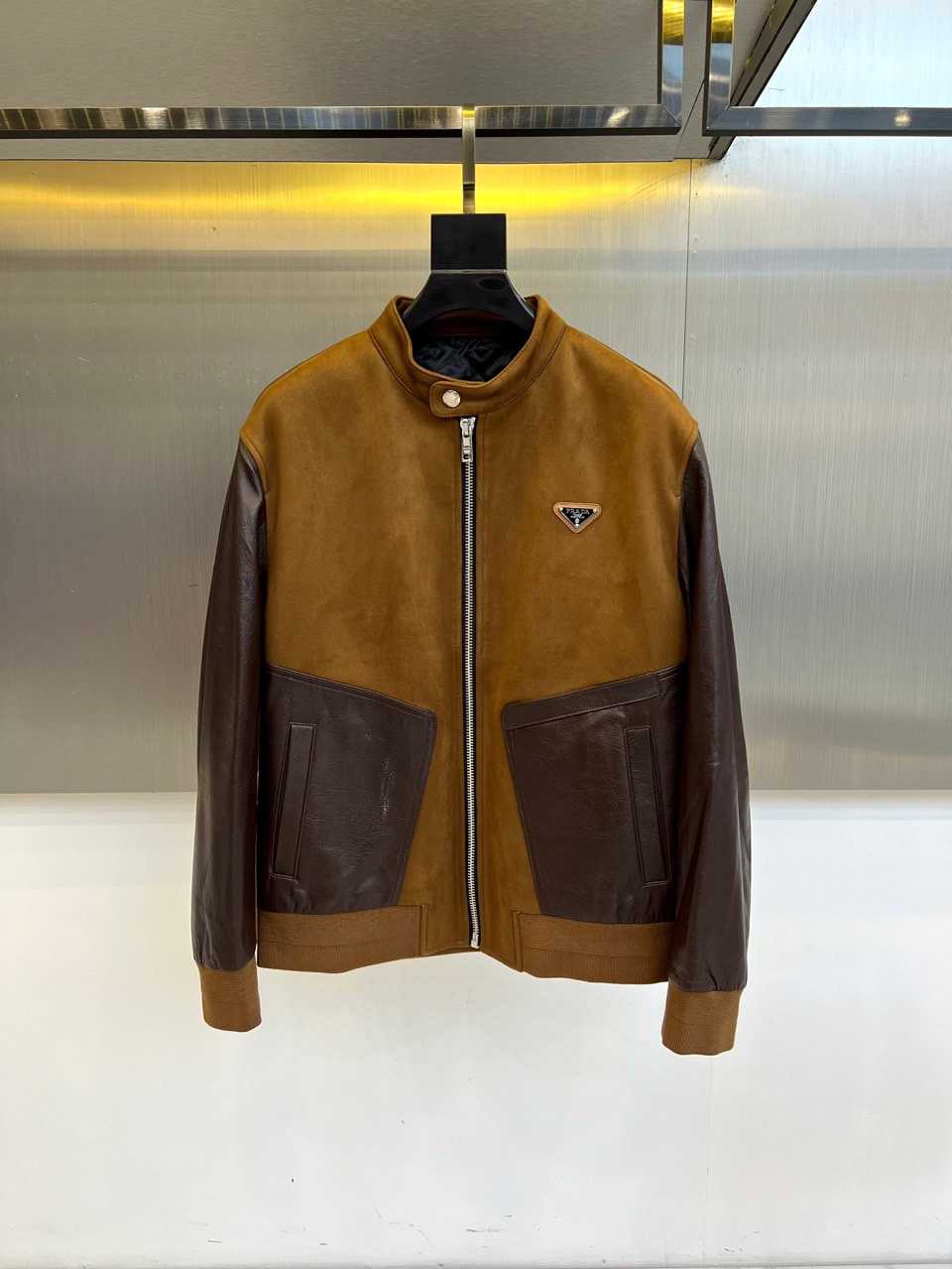 NO:681029,Prad, new products for autumn and winter  PRADA suede splicing jacket. This jacket is based on retro and modern design, with a stand-up collar design and a metal zipper opening and closing system. The overall body is made of brown suede material. The sleeves and side pockets are innovatively integrated into dark brown glossy leather splicing, forming a layered collision of texture and color. The left chest is embellished with the brand's classic triangular metal logo, continuing the iconic aesthetic gene. The soft touch of suede contrasts with the tough luster of the leather, making it neat and tidy.  The tailoring takes into account both self-cultivation and freedom of movement. It is suitable for a variety of wearing scenarios. It can be paired with casual pants to create an urban strolling look, or it can be paired with formal pants to demonstrate a business casual style. Through material splicing and detailed craftsmanship, PRADA's design philosophy of integrating Italian heritage and modern wearability is interpreted. 48-56 (model is 173, 143 pounds, upper body size 50),, prada, prada, jackets  & coats, chamois leather,19860909Prad普拉、秋冬新品 PRADA麂皮拼接夹克.这款夹克以复古摩登为设计基调,采用立领设计搭配金属拉链开合系统,整体以棕色麂皮材质构筑主体,袖身与侧袋创新融入深棕色光面皮革拼接,形成质感与色彩的层次碰撞.左胸点缀品牌经典三角形金属徽标,延续标志性美学基因.麂皮的柔软触感与皮革的硬朗光泽相映成趣,利落剪裁兼顾修身性与活动自由度.搭配适配多种穿搭场景,既可搭配休闲裤打造都市漫步造型,亦能配合正装裤演绎商务休闲风格.通过材质拼接与细节工艺,诠释PRADA将意式传承与现代实穿性相融合的设计哲学.48-56（模特173,143斤上身50码）,,prada,prada,jackets & coats,chamois leather,,Men's clothing