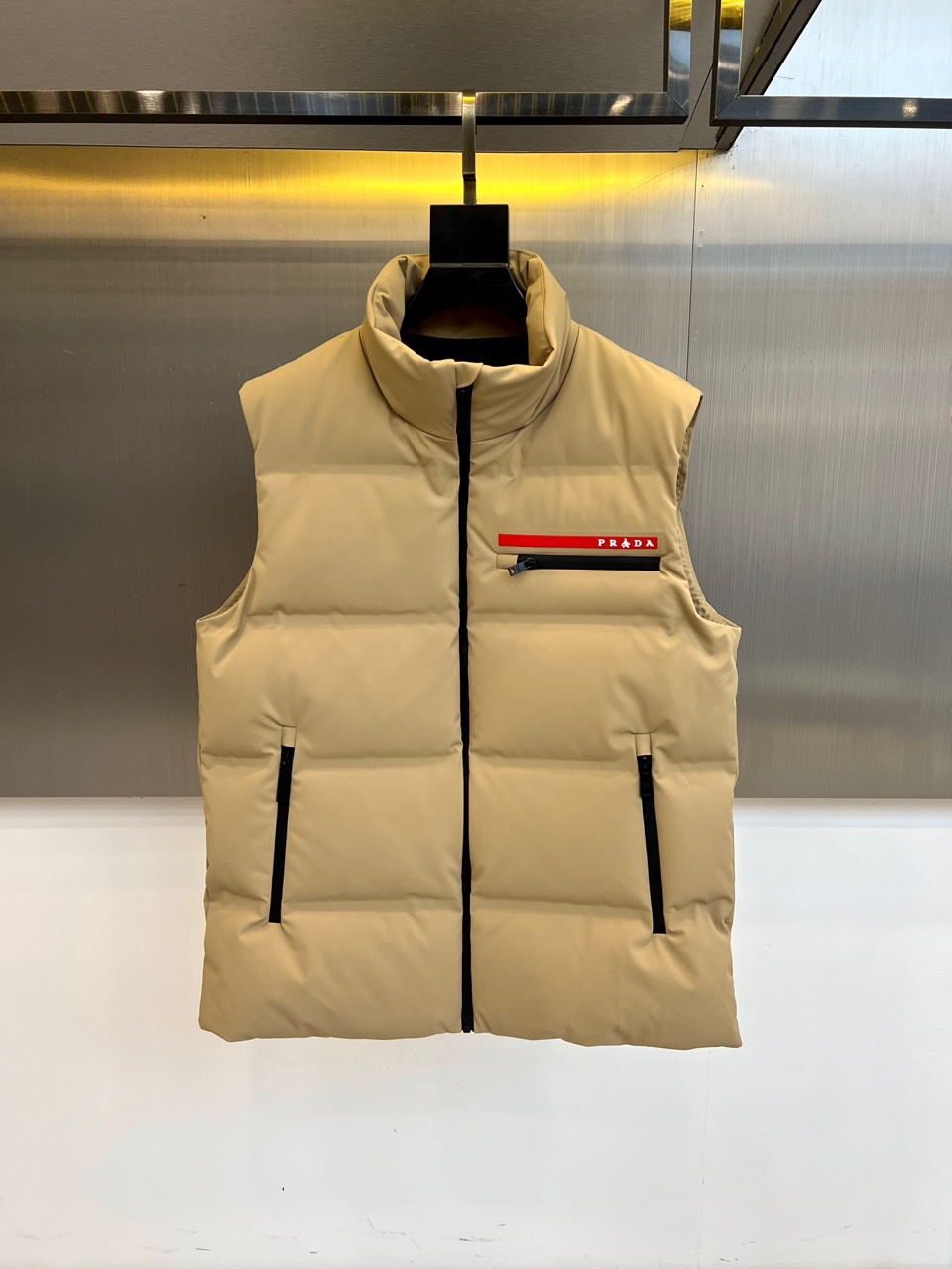 NO:681027,Prad, new product for autumn and winter, PRADA functional down vest. Stand-up collar design with sleeveless tailoring. It is equipped with a black smooth zipper closure system on the front and diagonal pockets with zippers on both sides. The chest is embellished with iconic patch pockets, with a red base and white PRADA words, and black and white stripes below. The fabric is blended with 87 nylon and 13 spandex, and the bottom layer is polyester fiber, filled with 90 down content white duck down, and the filling capacity is 110 grams.  The stand-up collar structure enhances wind resistance, and the red logo bag becomes the visual focus. It can be worn inside with a sweater to create a layered look, or worn outside to cope with slightly cold weather. Through the combination of practical details and brand elements, it interprets the design concept of integrating Italian aesthetics into functional clothing. 48-56 (model 173, 143 pounds, upper body size 50),, prada, prada, down jacket,19860909Prad普拉达、秋冬新品 PRADA机能风羽绒马甲. 立领设计搭配无袖剪裁,正面配置黑色顺滑拉链开合系统,两侧设带拉链的斜插口袋.胸部点缀标志性贴袋,以红色为基底搭配白色PRADA字样,下方饰有黑、白双色条纹.面料采用87锦纶与13氨纶混纺,底层为聚酯纤维,填充90绒子含量白鸭绒,充绒量达110克. 立领结构增强防风性,红色标识袋成为视觉焦点.可内搭针织衫打造层次造型,或外穿应对微寒天气.通过实用细节与品牌元素的结合,诠释将意式美学融入机能着装的设计理念.48-56（模特173,143斤上身50码）,,prada,prada,down jacket,,Men's clothing