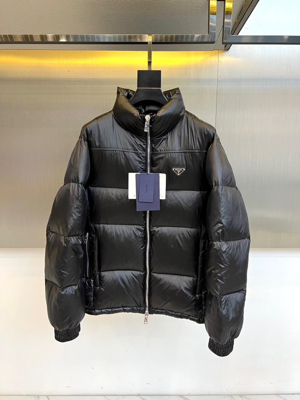 NO:681040,Prada, autumn and winter down jackets, reshape winter protection with minimalist aesthetics. The style adopts a neat stand-up collar and a smooth box-shaped silhouette, which is divided by precise horizontal quilting to outline geometric blocks with architectural layers. The iconic inverted triangle metal logo is embellished on the left chest, becoming a low-key and eye-catching brand imprint. The entire zipper closure, combined with the windproof design of the cuffs, combines simple styling with practical functions.  Its style is a rational expression of modern urban elites. The deep black and glossy eco-friendly fabrics highlight the low-key luxury texture. The core technology demonstrates the commitment to sustainability. The exterior and lining are made of recycled nylon, and the inner liner is recycled polyester fiber. It is filled with 90% high-quality white goose down, and the down filling amount reaches 161 grams, ensuring light, full and excellent warmth retention.  This down jacket deeply integrates Prada's pioneering design language and environmental protection concepts. It can not only cope with severe cold weather, but also perfectly integrate into urban daily life and business scenes. It is a model item for shaping a modern style and practicing responsible choices. 48-56 (model is 173, 143 pounds, upper body size 50),, prada, prada, down jacket,19860909Prad普拉达、秋冬羽绒服,以极简美学重塑冬日防护.款式采用利落立领与流畅的箱型廓形,通过精准的横向绗缝分割,勾勒出富有建筑层次的几何块面.标志性倒三角形金属徽标点缀于左胸,成为低调而醒目的品牌印记.全件拉链闭合,配合袖口防风设计,兼具简约造型与实用功能. 其风格是现代都市精英的理性表达.深邃的黑色与富有光泽的环保面料,凸显低调奢华的质感.核心技术彰显可持续承诺外部及衬里均采用再生锦纶,内胆为再生聚酯纤维.填充90高品质白鹅绒,充绒量达161克,确保轻盈饱满的卓越保暖性. 这件羽绒服将普拉达的先锋设计语言与环保理念深度融合,既能从容应对严寒气候,亦能完美融入都市日常与商务场景,是塑造摩登型格与践行责任选择的典范单品.48-56（模特173,143斤上身50码）,,prada,prada,down jacket,,Men's clothing