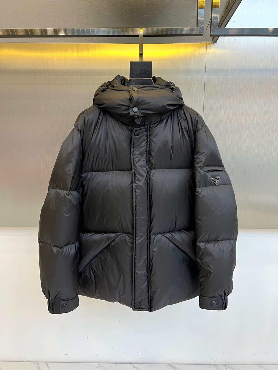 NO:681052,Prada, the autumn and winter black down jacket, reshapes the warmth of winter with its iconic design language. The style adopts a loose and casual hooded silhouette, and the brim is equipped with adjustable buttons to enhance practical details. The entire body of the garment is outlined with regular block seams, forming an architectural three-dimensional silhouette. The front is simple zipper closure, and the left sleeve is embellished with a classic triangular metal logo, low-key declaring the brand identity.  Its style is an exquisite fusion of modern minimalism and street casualness. The pure black color and loose fit create a casual and uninhibited fashion attitude. It can be paired with jeans and sneakers for a relaxed daily life; it can also be worn with a simple skirt to create a different kind of sophistication. The excellent warmth is hidden in the crisp shape, making it a popular choice for autumn and winter.  A practical choice that combines style and warmth. This down jacket is not only a cold-proof item, but also an essential equipment that shows low-key luxury and contemporary style. 48-56 (model weighs 173,143 pounds, upper body size 50),, prada, prada, sneakers, skirts, jeans, down  jacket,hats,19860909Prad普拉达、秋冬黑色羽绒服,以标志性设计语言重塑冬日温暖.款式采用宽松休闲的连帽廓形,帽檐配有可调节纽扣,提升实用细节.衣身通体以规整的块状衍缝勾勒,形成富有建筑感的立体轮廓.正面简洁的拉链开合,左袖点缀经典的三角形金属徽标,低调宣告品牌身份. 其风格是现代极简主义与街头休闲感的精妙融合.纯粹的黑色与宽松版型,营造出随性不羁的时髦态度.它既能与牛仔裤和运动鞋搭配,演绎轻松日常；也可内搭简约裙装,碰撞出别样的轻熟优雅.卓越的保暖性隐含于挺括造型之中,成为秋冬兼具风度与温度的实用之选.这件羽绒服不仅是御寒单品,更是彰显低调奢华与当代型格的必备装备.48-56（模特173,143斤上身50码）,,prada,prada,sneakers,skirts,jeans,down jacket,hats,,Men's clothing