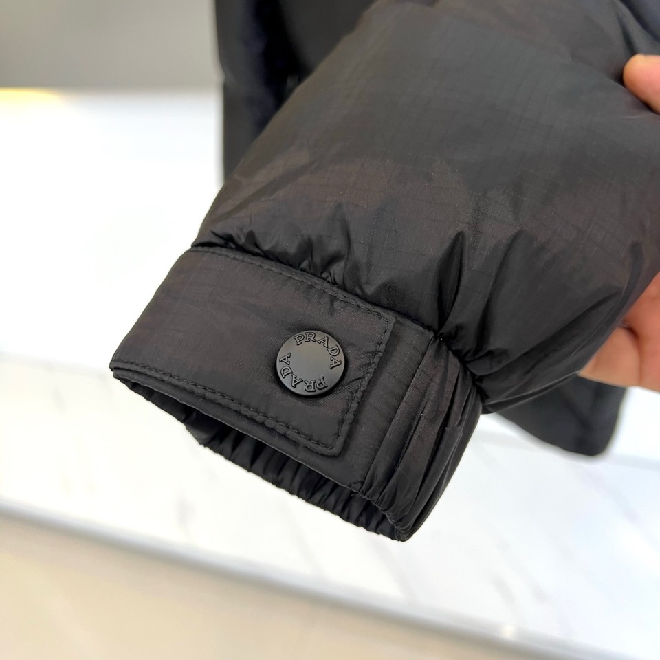 Prad普拉达、2025秋冬新品黑色羽绒服，以经典黑色重塑都市冬日美学。款式采用简约连帽设计，帽部点缀实
