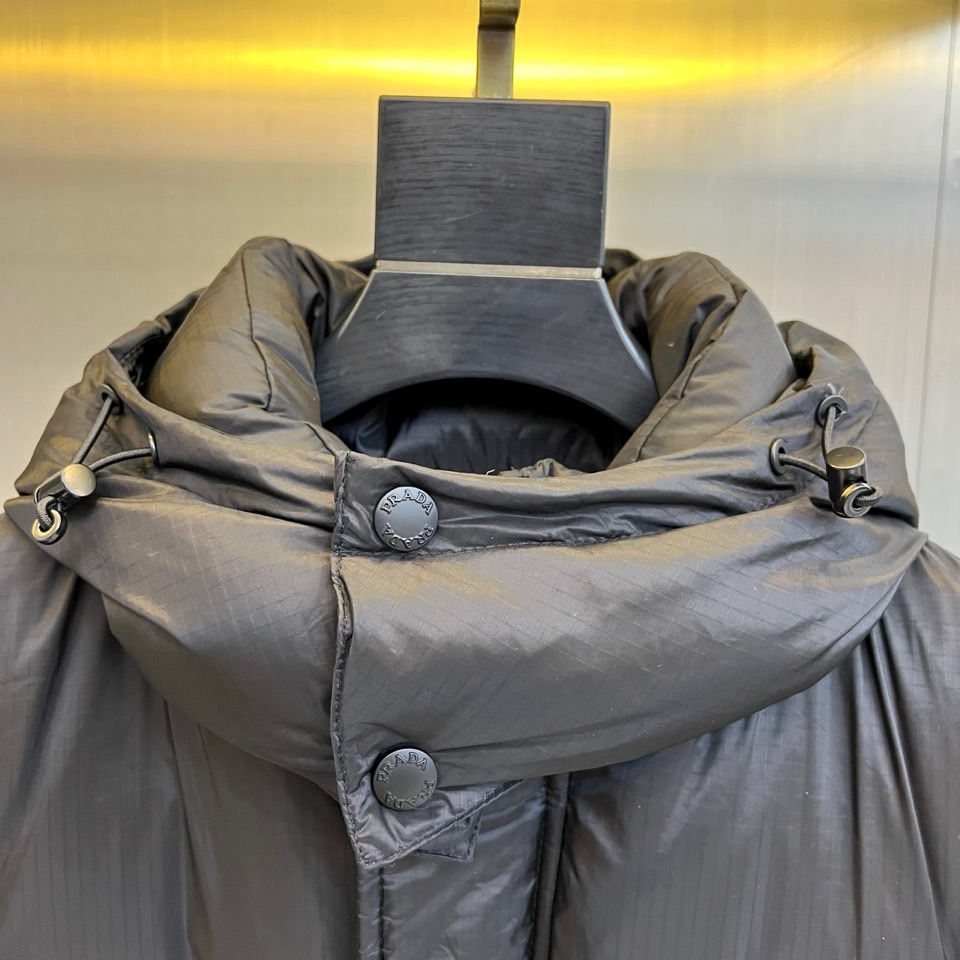 Prad普拉达、2025秋冬新品黑色羽绒服，以经典黑色重塑都市冬日美学。款式采用简约连帽设计，帽部点缀实