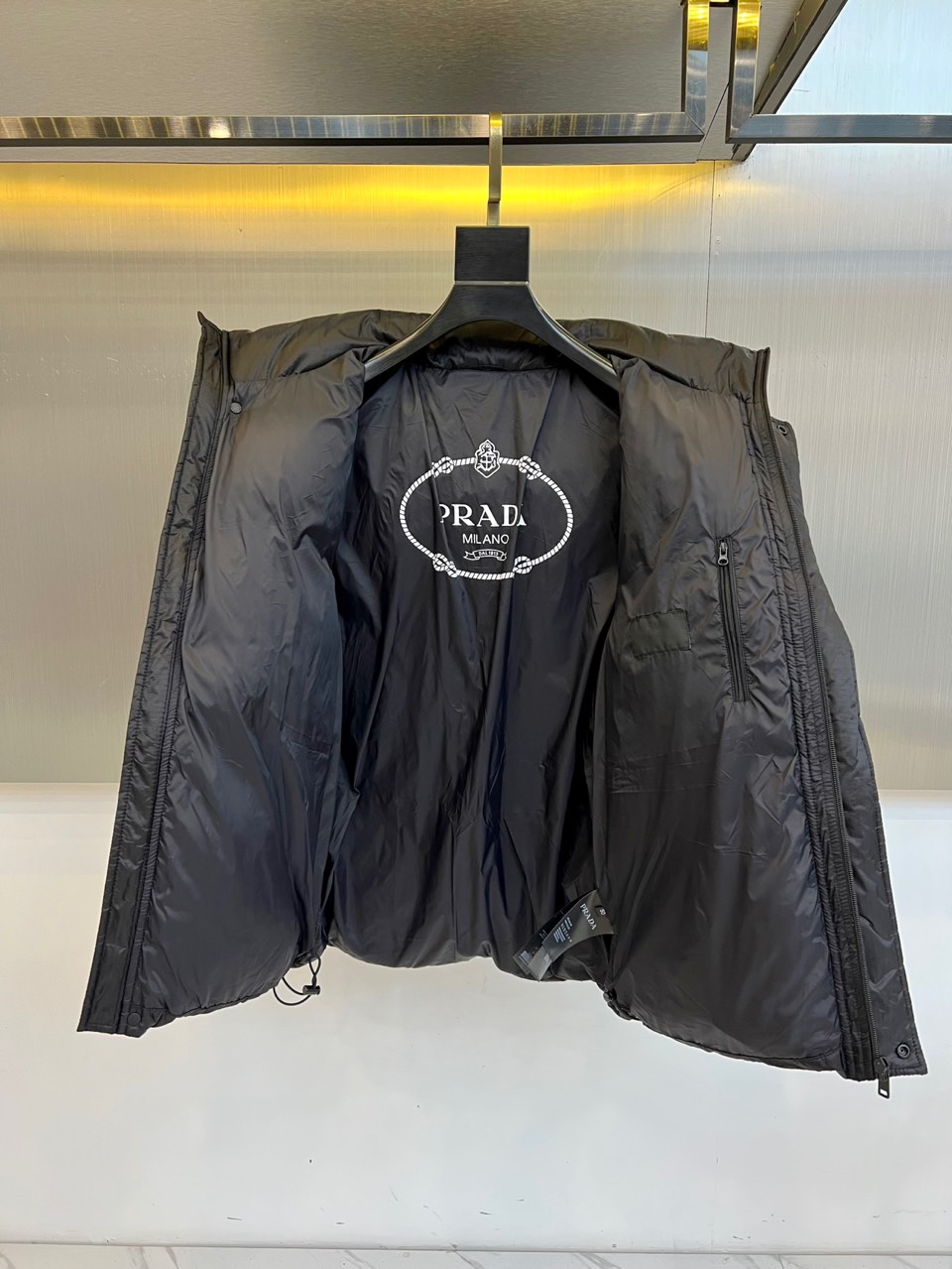 Prad普拉达、2025秋冬新品黑色羽绒服，以经典黑色重塑都市冬日美学。款式采用简约连帽设计，帽部点缀实