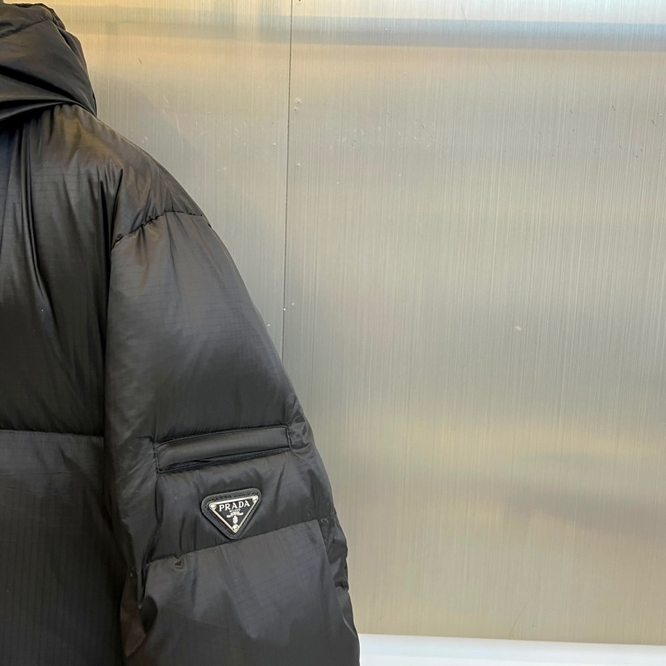 Prad普拉达、2025秋冬新品黑色羽绒服，以经典黑色重塑都市冬日美学。款式采用简约连帽设计，帽部点缀实