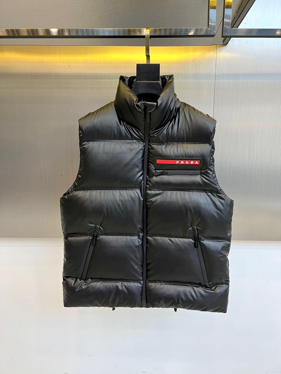 NO:712025,Prada's 2025 autumn and winter new down vest interprets modern minimalist aesthetics with a pure black tone. The style adopts a neat stand-up collar design, with a smooth zipper opening and closing system. The body is covered with regular diamond quilting texture, and the three-dimensional filling process ensures that 90 high-quality white duck down is evenly distributed. It is equipped with practical pockets with zippers on both sides, and the left chest is embellished with the iconic red webbing label, and the white PRADA words highlight the brand genes. The fabric is made of recycled fiber material, reflecting the concept of environmental protection and sustainable fashion proposition.  The style is a perfect fusion of Italian rationalism and practical philosophy. The slim tailoring maintains warmth without losing the sense of lightness. The all-black color scheme is both calm and versatile. It can be worn with a sweater to create a layered look or with a coat to enhance the overall fashion. From environmentally friendly fabrics to precise quilting technology, every detail  All reflect Prada's insistence on quality and responsibility. This down vest is not only a practical tool for layering in autumn and winter, but also interprets the sustainable luxury attitude of modern wardrobes with low-key details. 48-56 (model weighs 173,143 pounds, upper body size 50),, prada, prada, down  jacket,19860909Prad普拉达、2025秋冬新品羽绒马甲,以纯黑色调诠释现代极简美学.款式采用利落立领设计,搭配顺滑拉链开合系统,衣身覆盖规整的菱形绗缝纹理,通过立体填充工艺确保90高品质白鸭绒均匀分布.两侧配备带拉链的实用插袋,左胸点缀标志性红色织带标签,白色PRADA字样凸显品牌基因.面料选用再生纤维材质,体现环保理念与可持续时尚主张. 风格上完美融合意式理性主义与实用哲学,修身剪裁既保持保暖性又不失轻盈感.全黑配色兼具沉稳与百搭特性,既能内搭针织衫打造层次造型,也可外搭大衣增强整体时尚度.从环保面料到精准绗缝工艺,每处细节都体现普拉达对品质与责任的坚持,这件羽绒马甲不仅是秋冬叠穿的实用利器,更以低调细节诠释现代衣橱的可持续奢华态度.48-56（模特173,143斤上身50码）,,prada,prada,down jacket,,Men's clothing